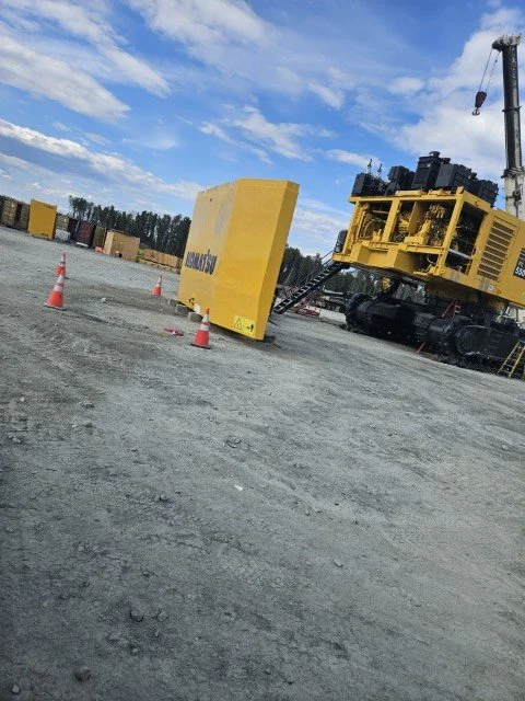 Construction site with large yellow machinery, safety cones, and storage containers in the background under a partly cloudy sky.