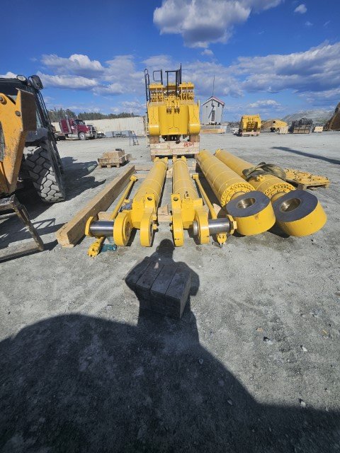 Construction equipment parts, including yellow rollers, beams, and a hydraulic lift, are arranged outdoors on gravel with vehicles and a building in the background.