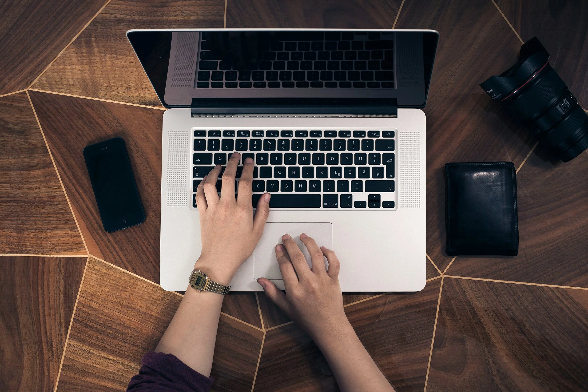 Top-down view of a person's hands operating a silver laptop on a wooden desk, with a smartphone, black wallet, and camera nearby.