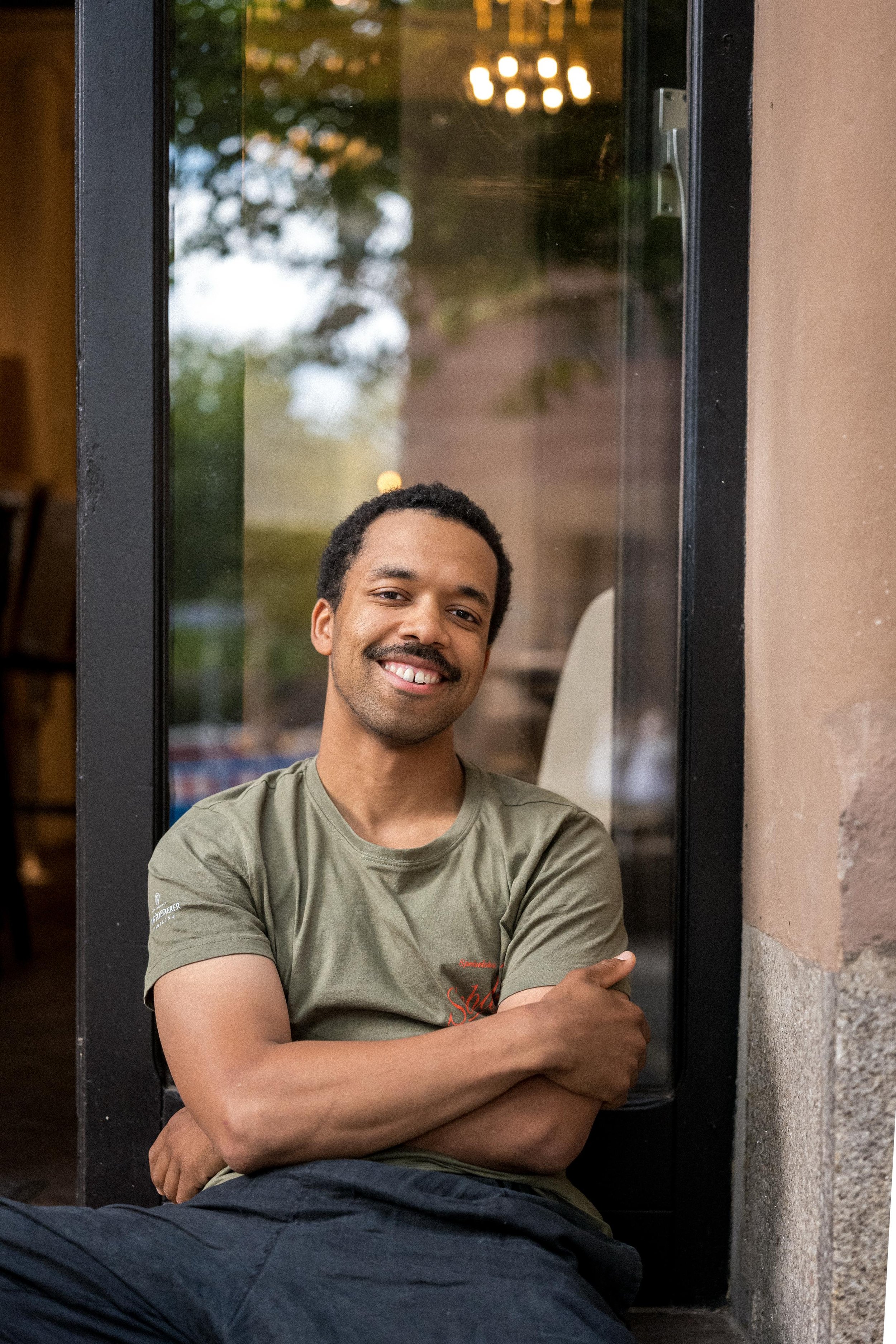 A young man with a mustache and beard smiling, sitting with arms crossed, leaning against a wall next to a large window with a black frame, showing a reflection of a chandelier and trees outside.