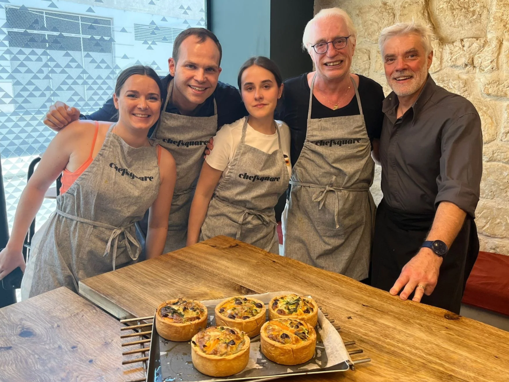 Six people, four women and two men, stand around a wooden table with freshly baked savory French vegetarian tourte. They are smiling and wearing aprons, in a kitchen setting with a stone wall and decorative window behind them.