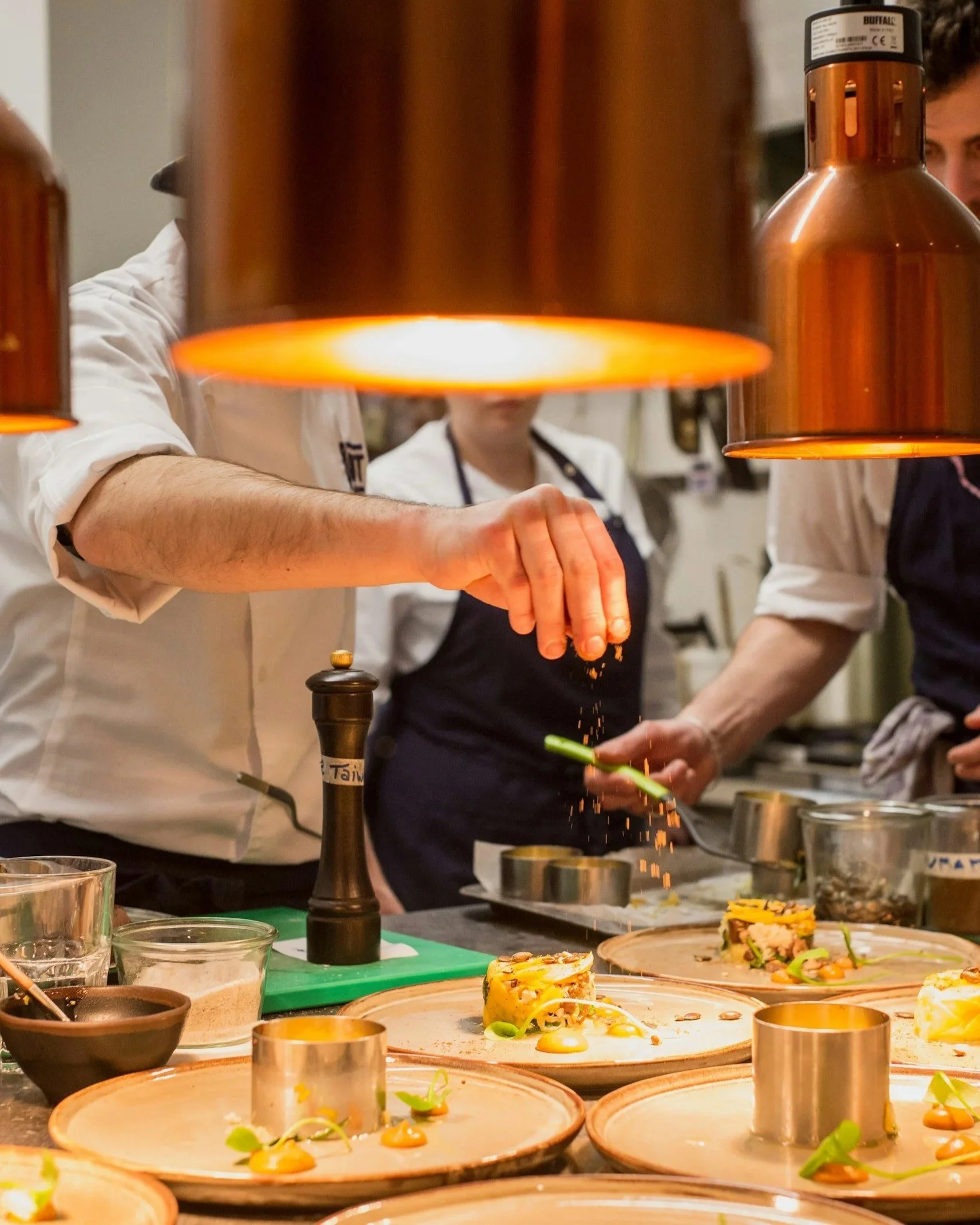 Chefs preparing plated dishes in a professional kitchen with warm lighting and multiple plates of food on the counter.
