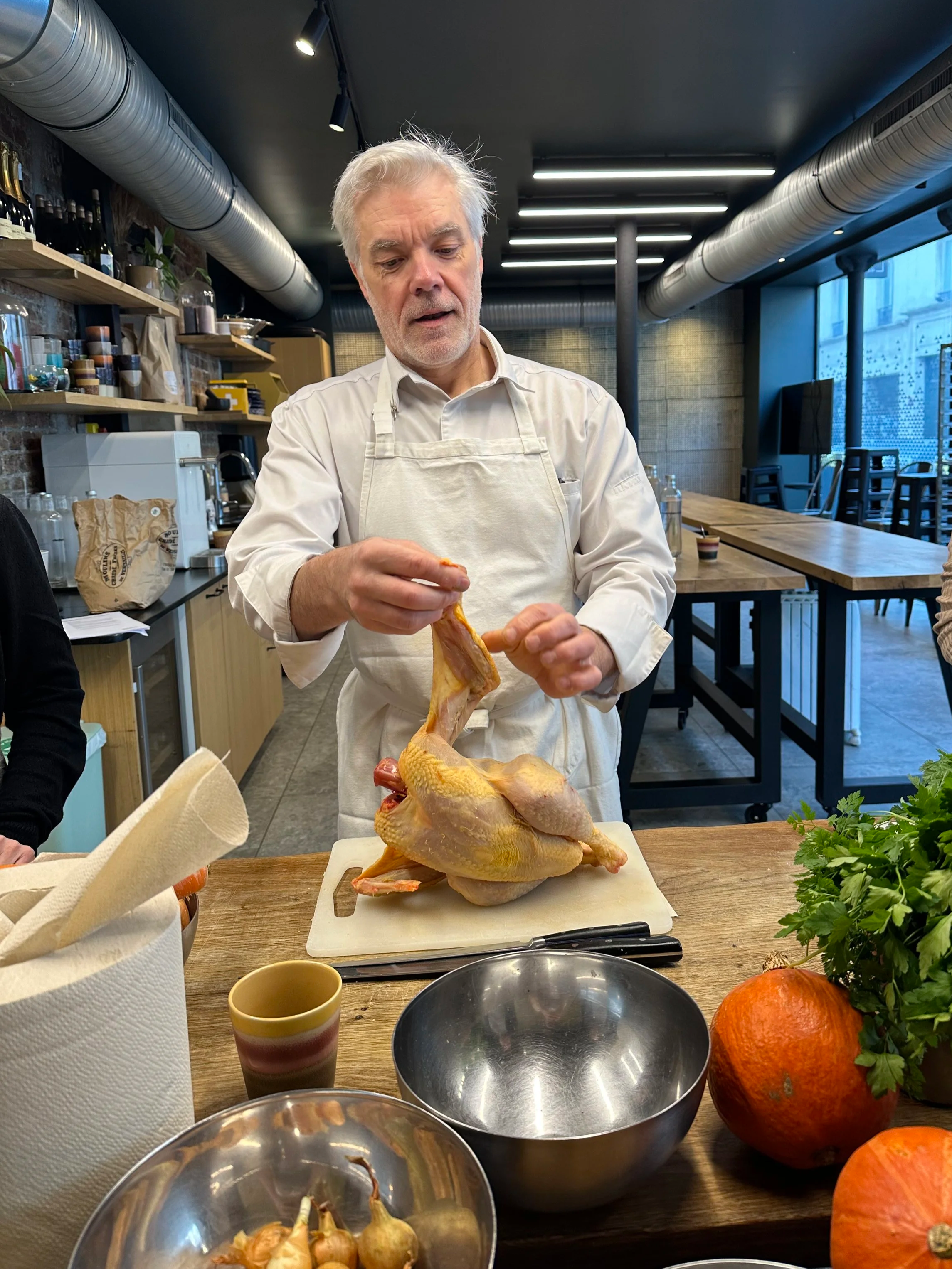 A man in a white apron is preparing a raw chicken on a cutting board in a modern kitchen or restaurant setting.
