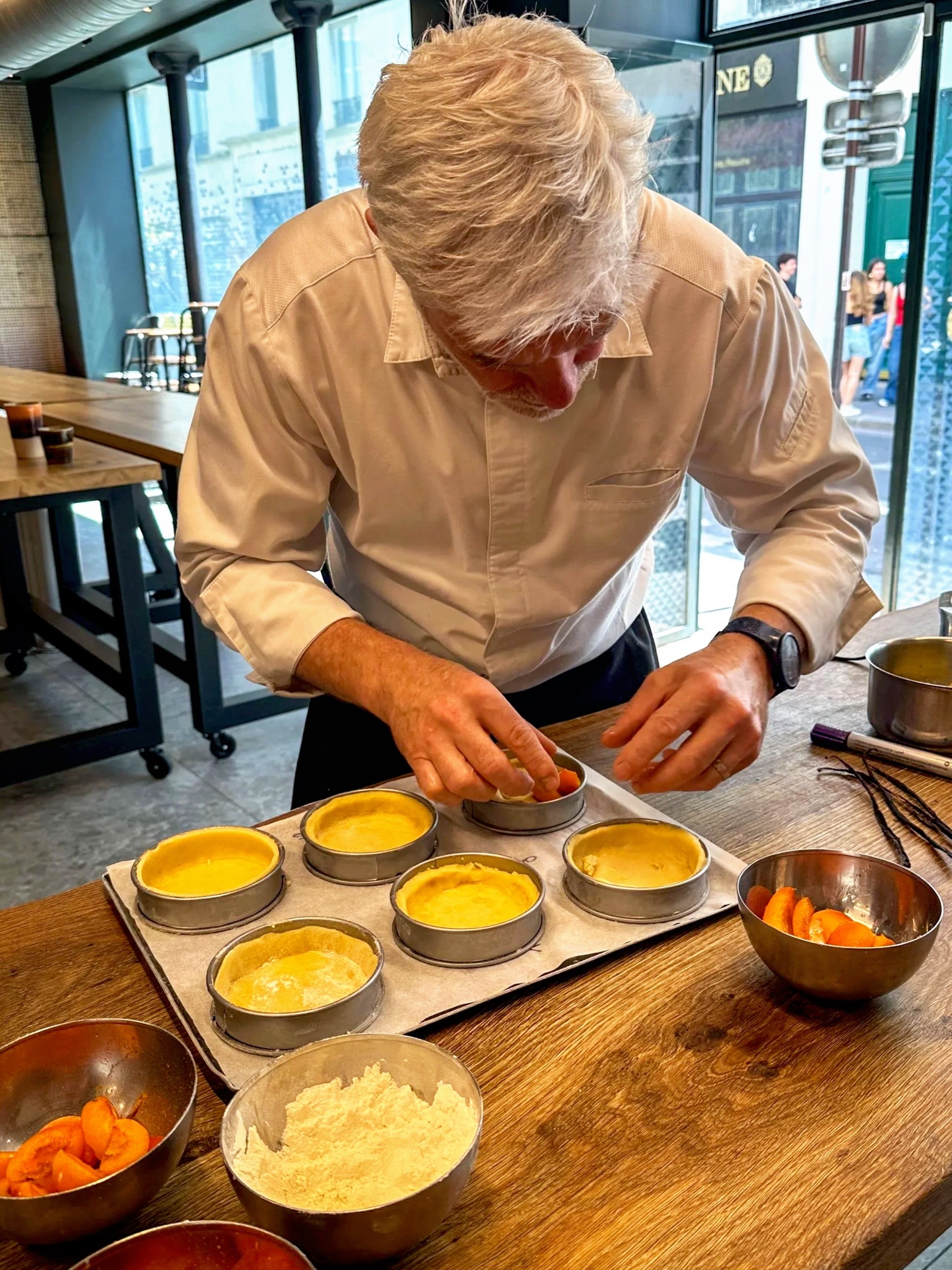 A man with white hair wearing a white shirt and a watch is preparing small desserts like apricot pie, in a cooking studio, surrounded by bowls of fruit and a tray of tart shells.