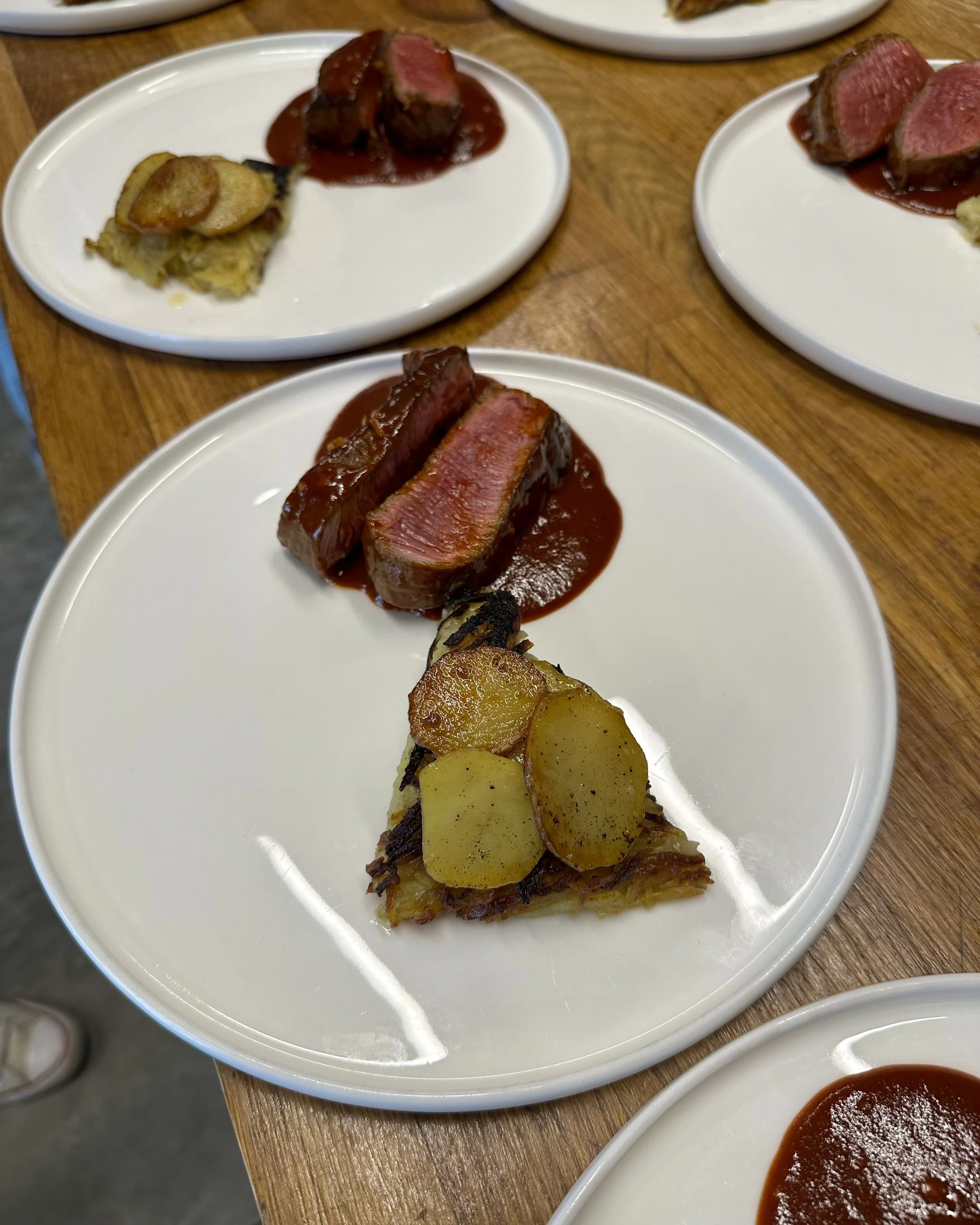 Three plates of sliced beef with red sauce, a piece of casserole with potatoes, and a slice of baked dish topped with potatoes on a wooden table.