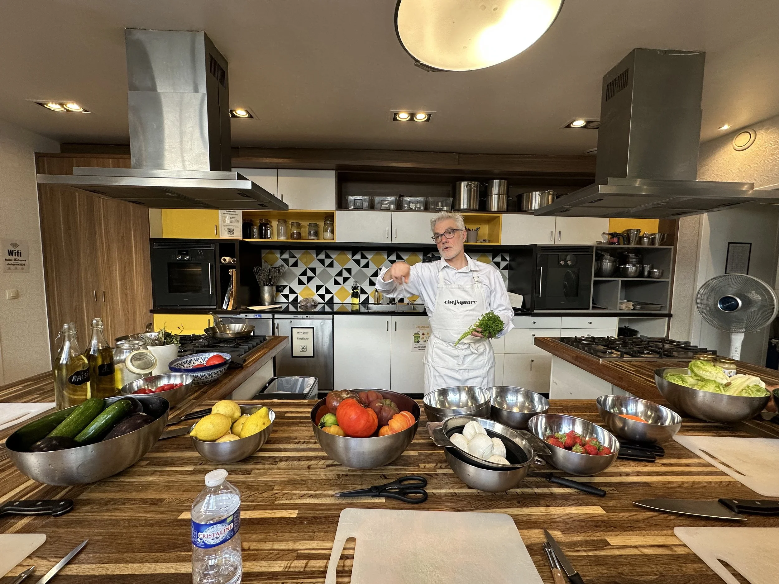 A man with gray hair and glasses wearing a white apron stands in a modern kitchen, holding a bunch of green parsley. The kitchen has a wooden countertop filled with bowls of vegetables including cucumbers, lemons, tomatoes, strawberries, and garlic. There are various kitchen utensils and appliances, with spice jars arranged on shelves and two large stainless steel range hoods above the cooking area. The background features a mix of yellow, black, white, and wooden cabinetry and a geometric backsplash. A water bottle is on the countertop in the foreground.