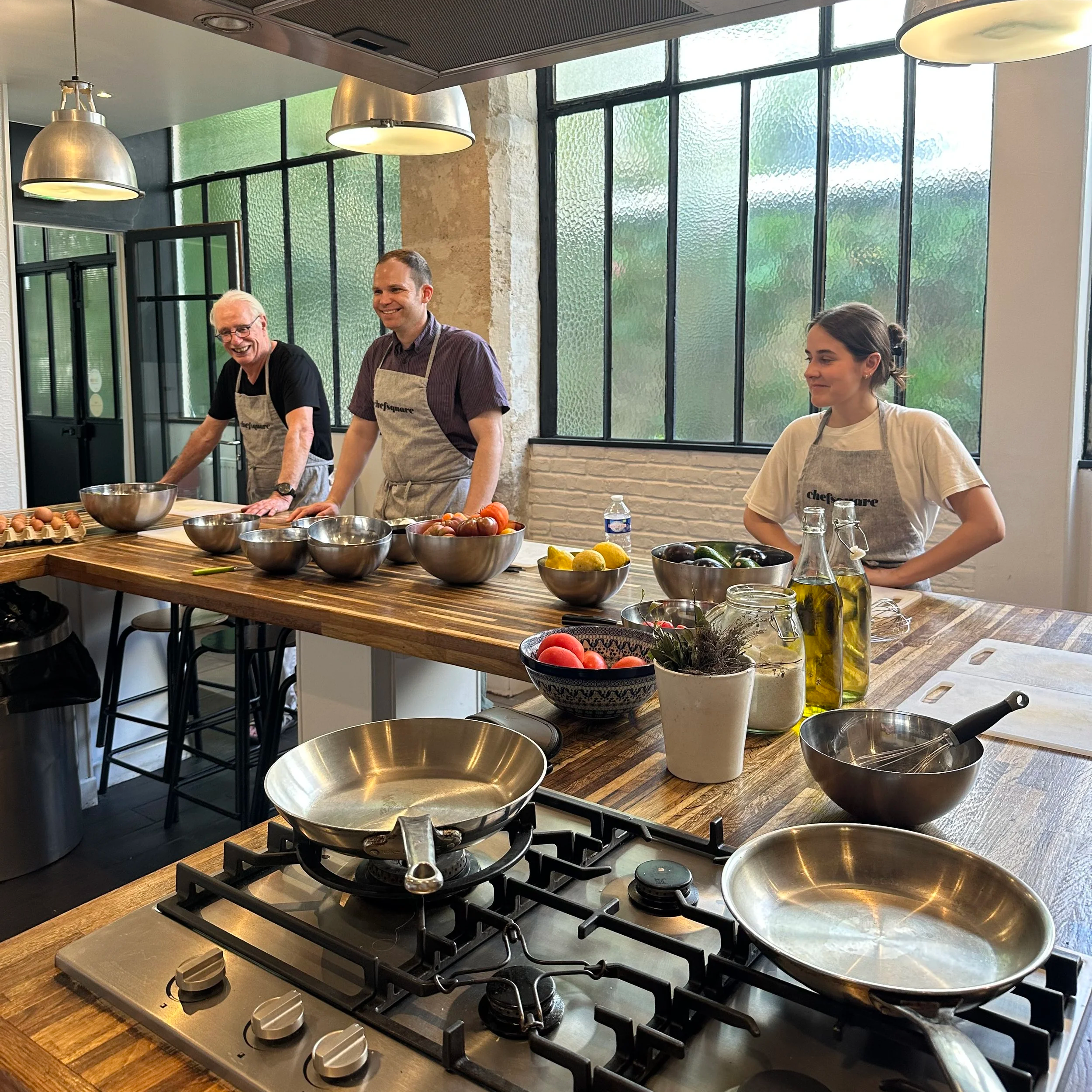 Three people smiling and standing behind a kitchen island with bowls of various ingredients, including eggs, tomatoes, and zucchini, in a well-lit, modern kitchen with large windows, a stainless steel stove with two pans, and kitchen utensils and bottles on the counter.