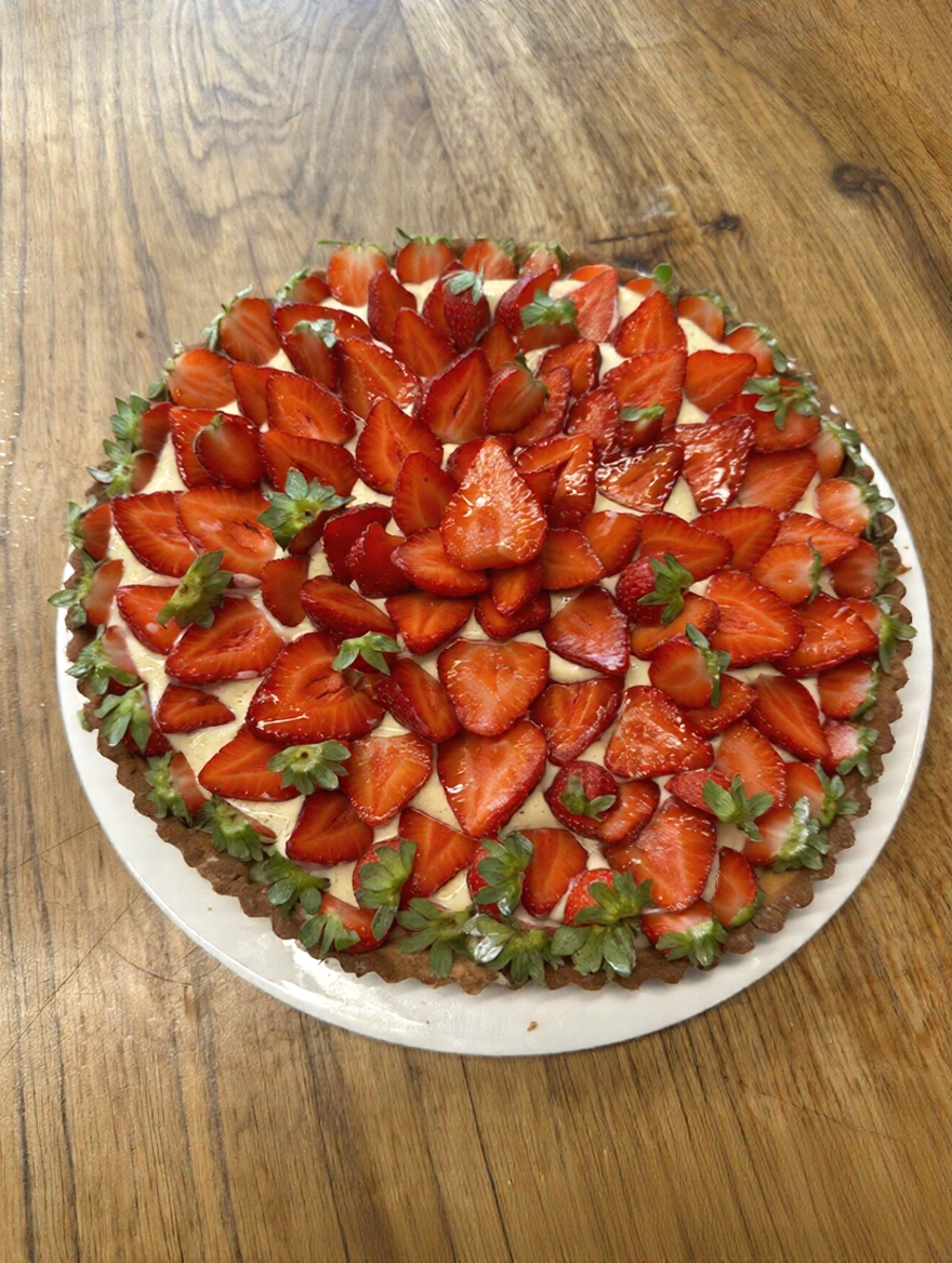 Strawberry tart with sliced strawberries on top of a creamy filling, decorated with whole and halved strawberries and green leaves, on a white plate on a wooden table.