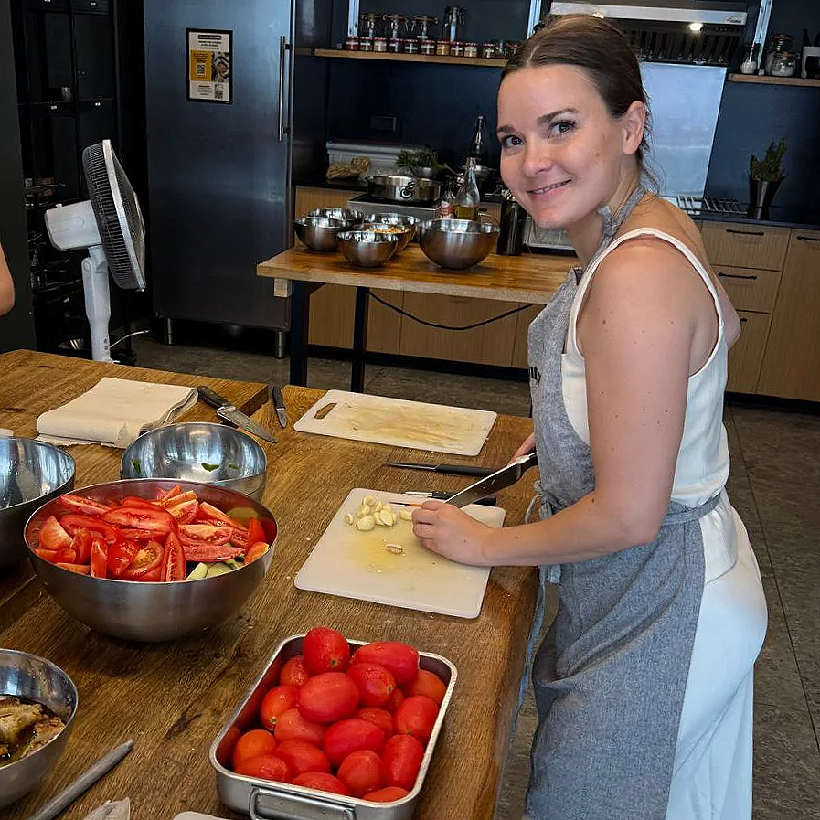 A woman wearing a grey apron is chopping garlic in a kitchen with a wooden countertop, surrounded by bowls of chopped tomatoes, whole tomatoes, and other cooking utensils.
