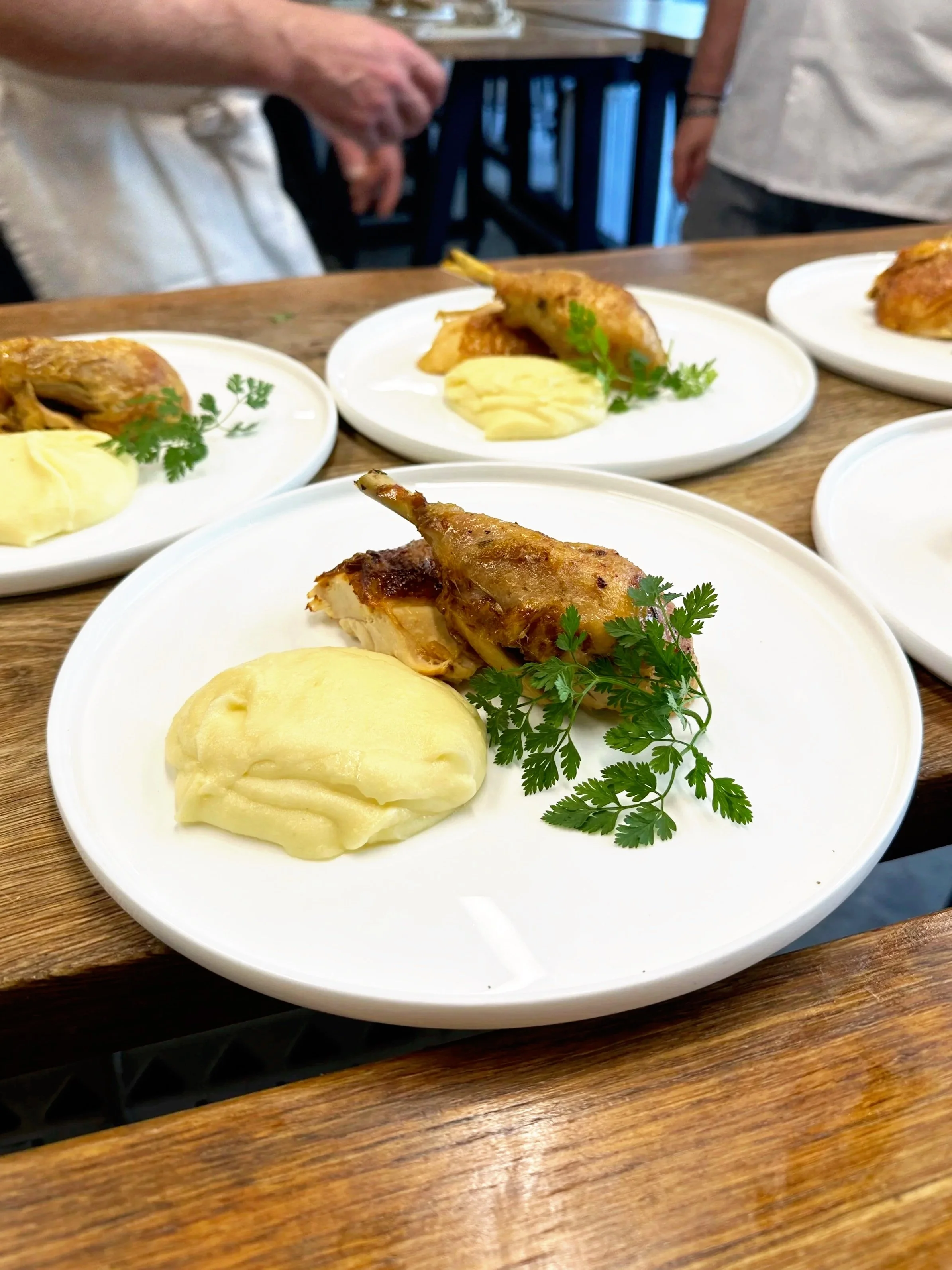 A white plate with potato puree, roasted chicken drumstick with herbs, and garnished with parsley. Multiple similar plates are on a wooden table in the background.
