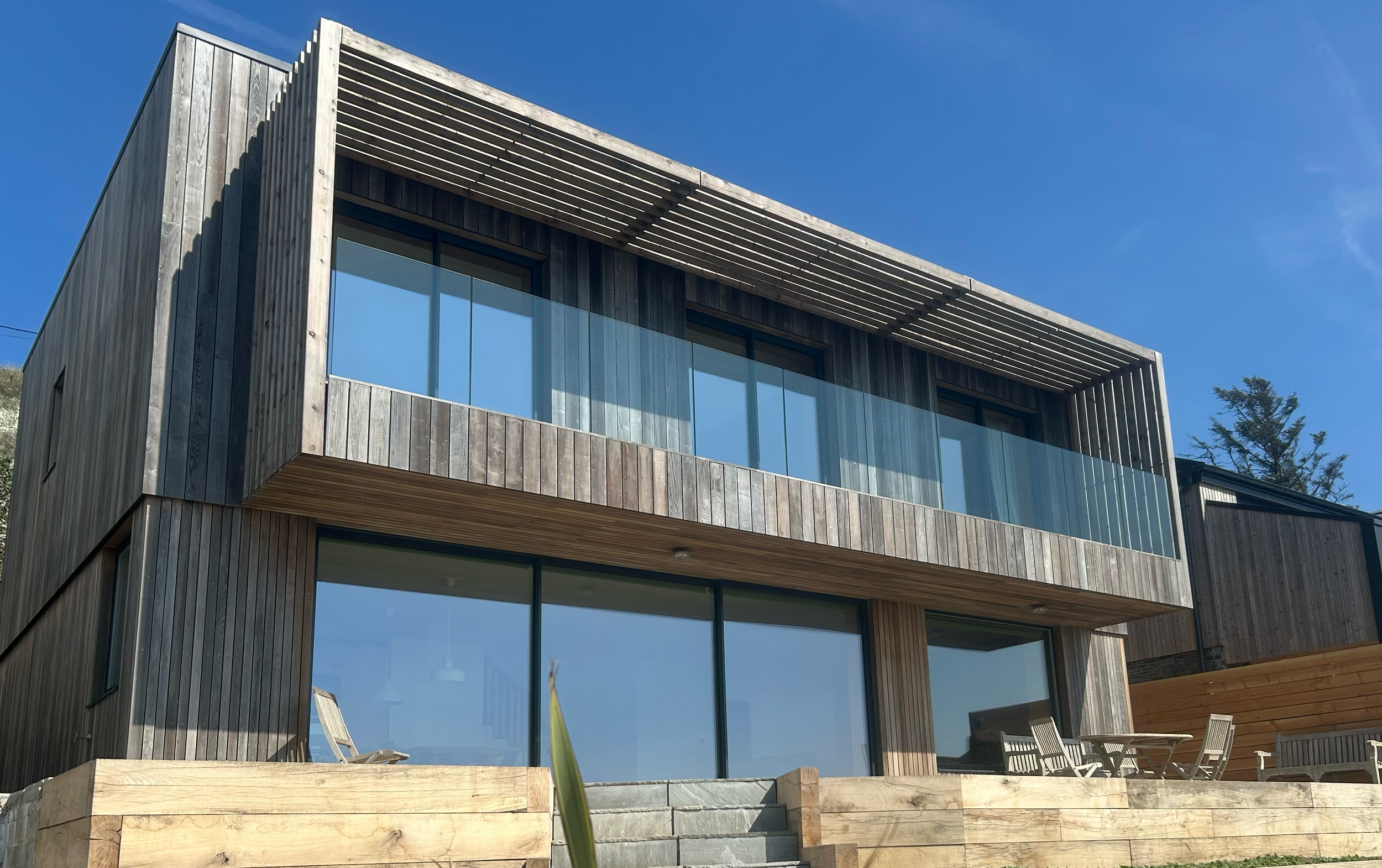 Modern multi-story house with wooden exterior and large glass windows, surrounded by a patio with outdoor furniture, under a bright blue sky. Timber frame property with cantilevered balcony.
