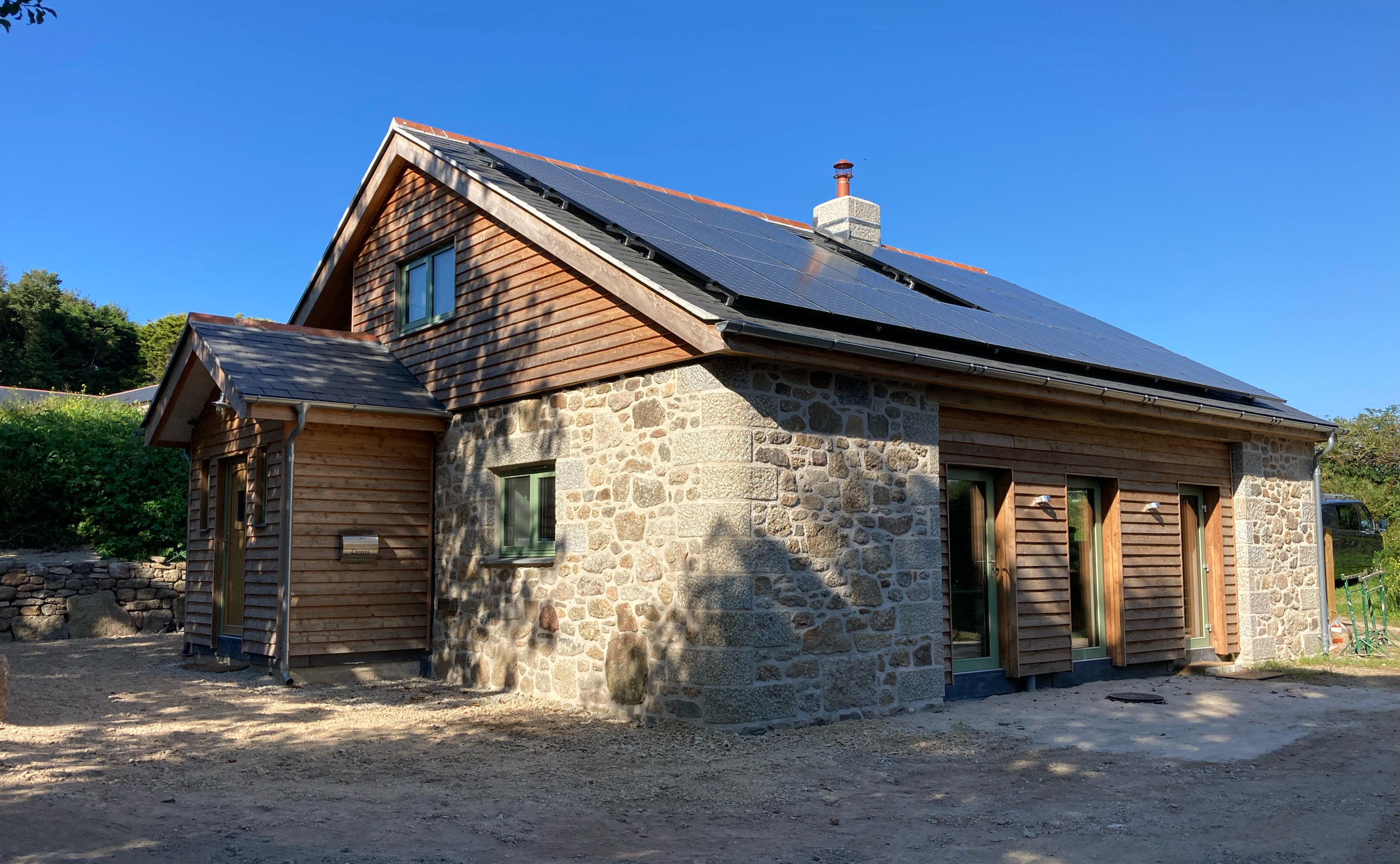 A house with a stone and wood exterior, and a roof with solar panels, under a clear blue sky. Terras won the Cornish building group award 2022.