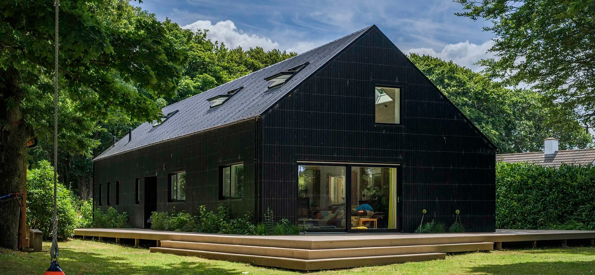 Black modern house with large windows and a sloped roof surrounded by green trees and lawn. Timber frame property with Black cladding.