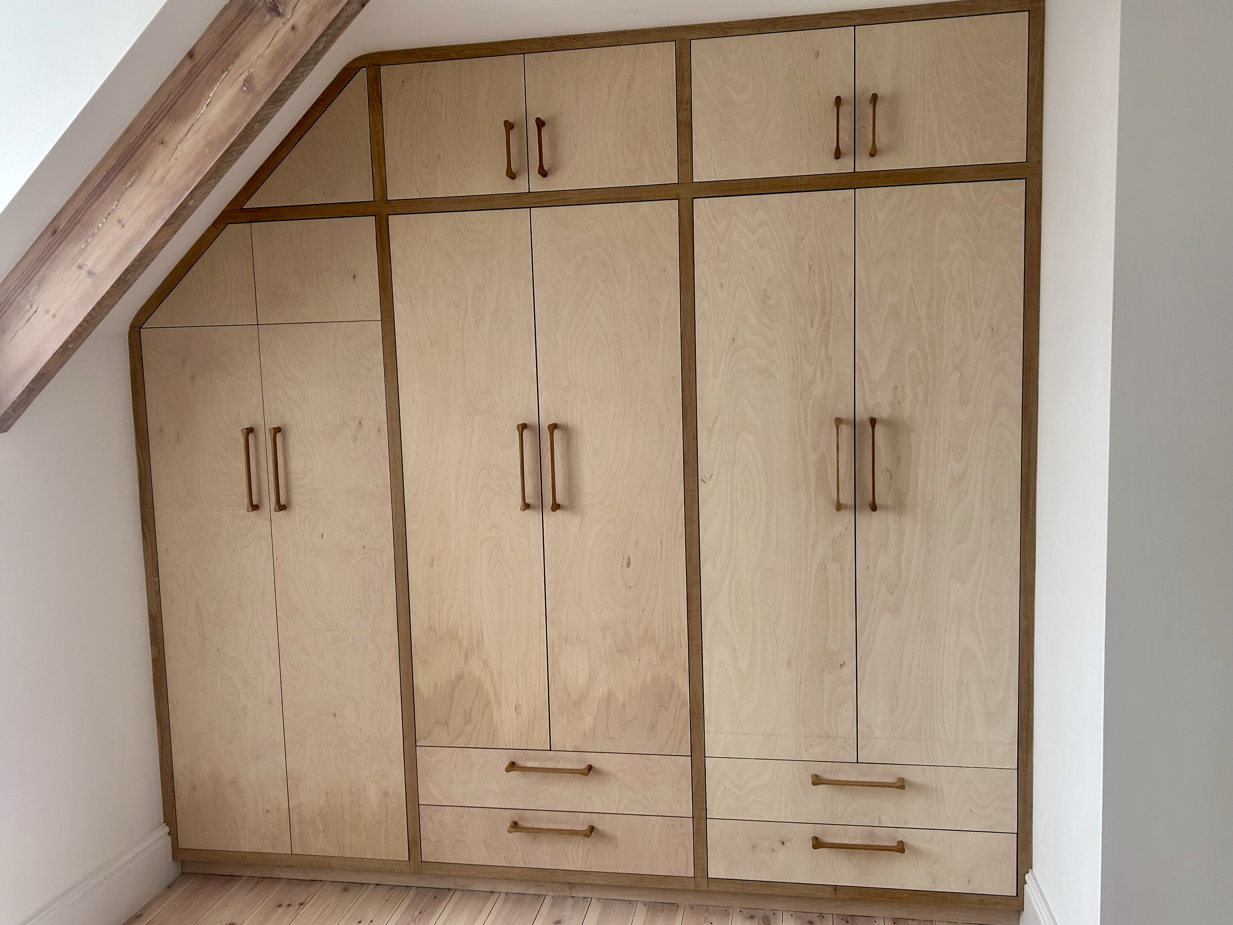Custom light Birch Ply built-in wardrobe with cupboards for hanging clothes, drawers, and wooden handles beneath a sloped ceiling