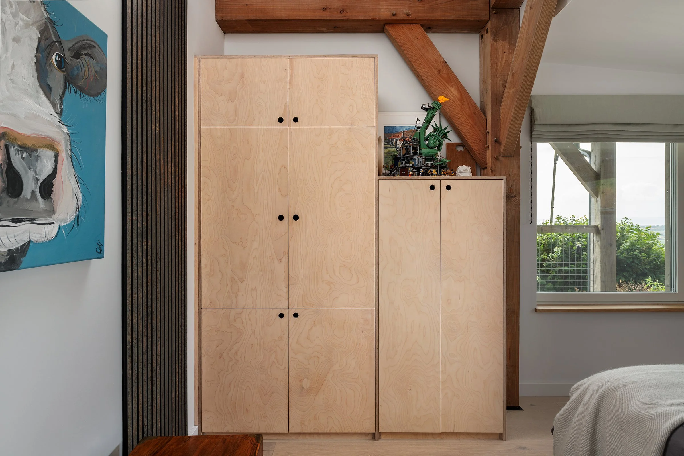 Modern bedroom with plywood storage cabinets, exposed timber beams, and window overlooking greenery