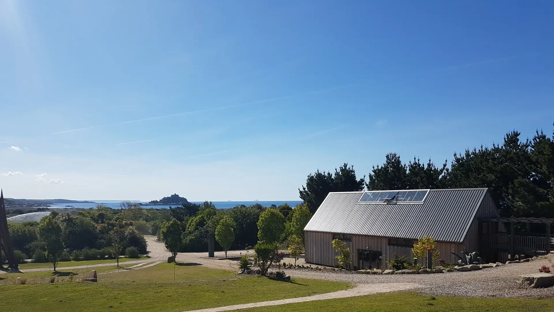 Scenic view of a coastal landscape with an island in the distance, a clear blue sky, a house with a metal roof and large windows, surrounded by trees and greenery. Purpose built gallery in Penzance Cornwall.