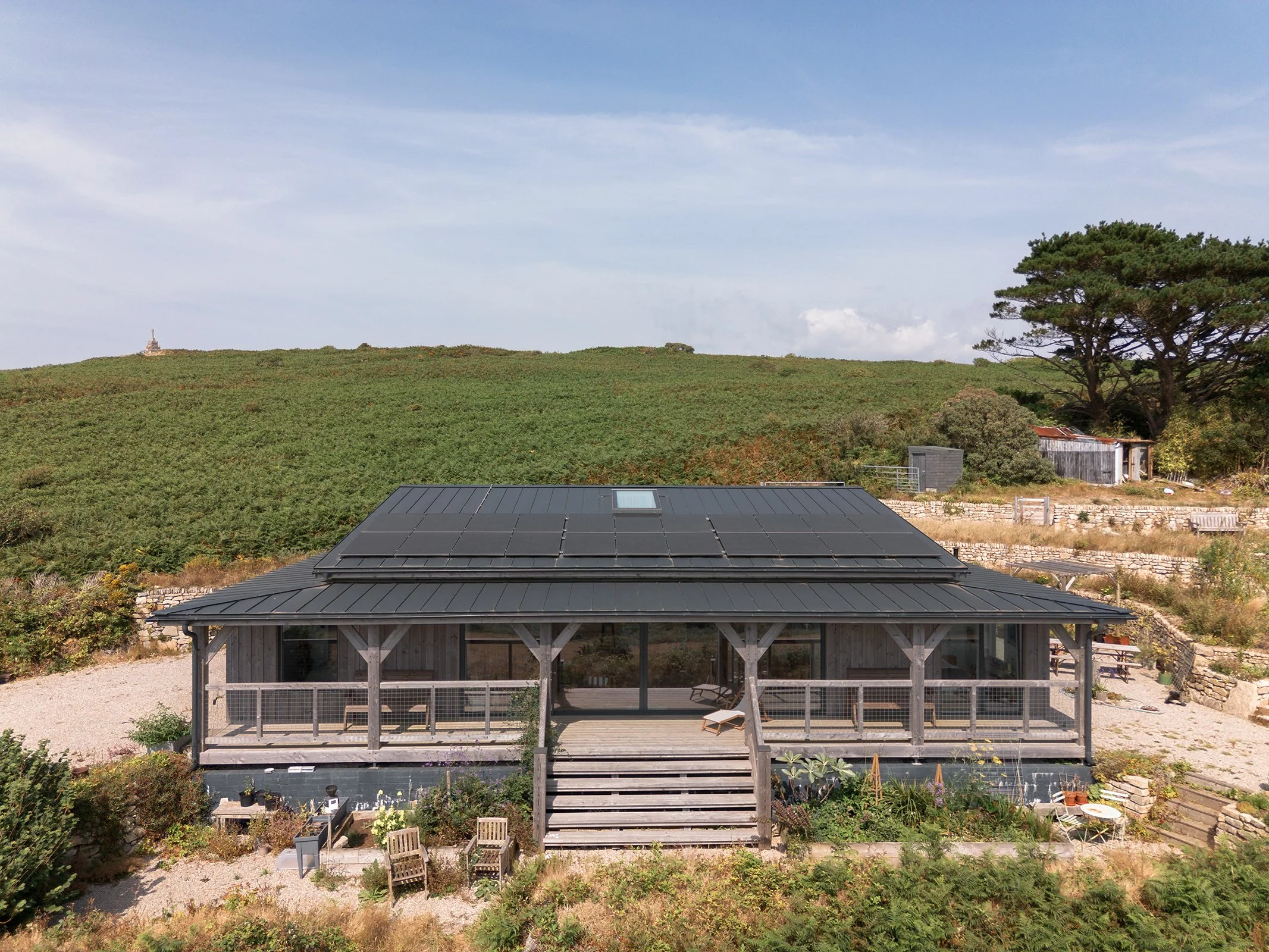 Skyfold, beautiful panoramic view, Timber frame property, surrounded by a garden and outdoor furniture, with a hill covered in greenery and a large tree in the background, under a blue sky with some clouds.