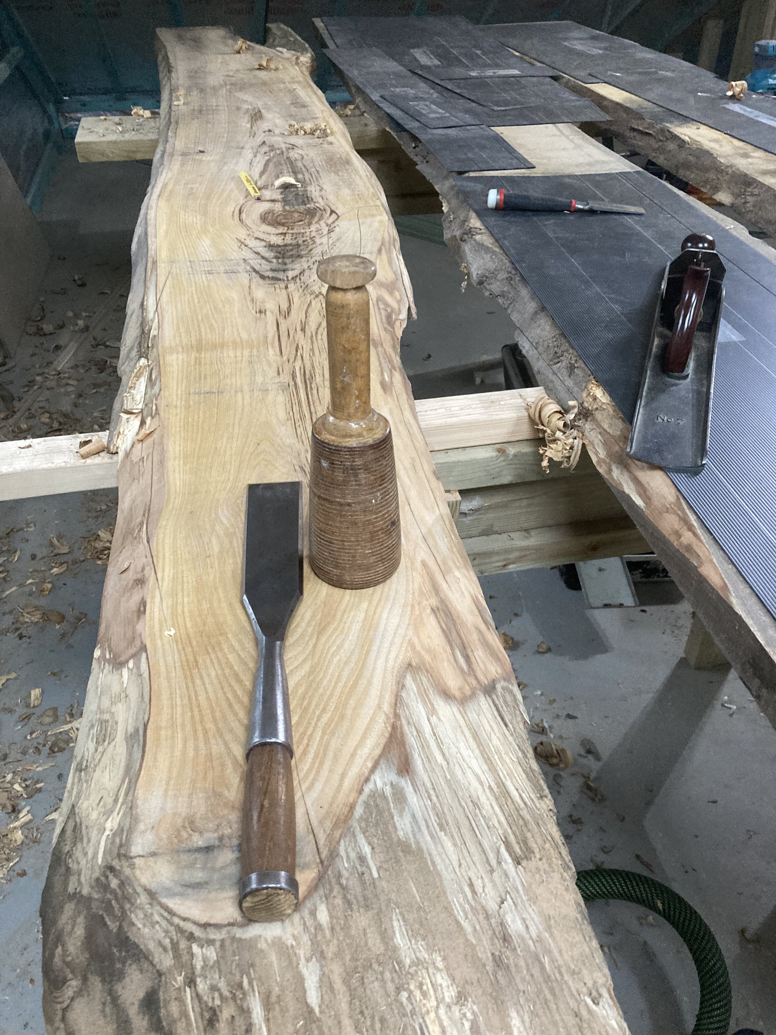 A woodworking workshop with a large piece of wood on a workbench, a chisel, a clamp, a mallet, and several pieces of sandpaper. Wood shavings and sawdust are scattered around. Craftsmanship at its best.