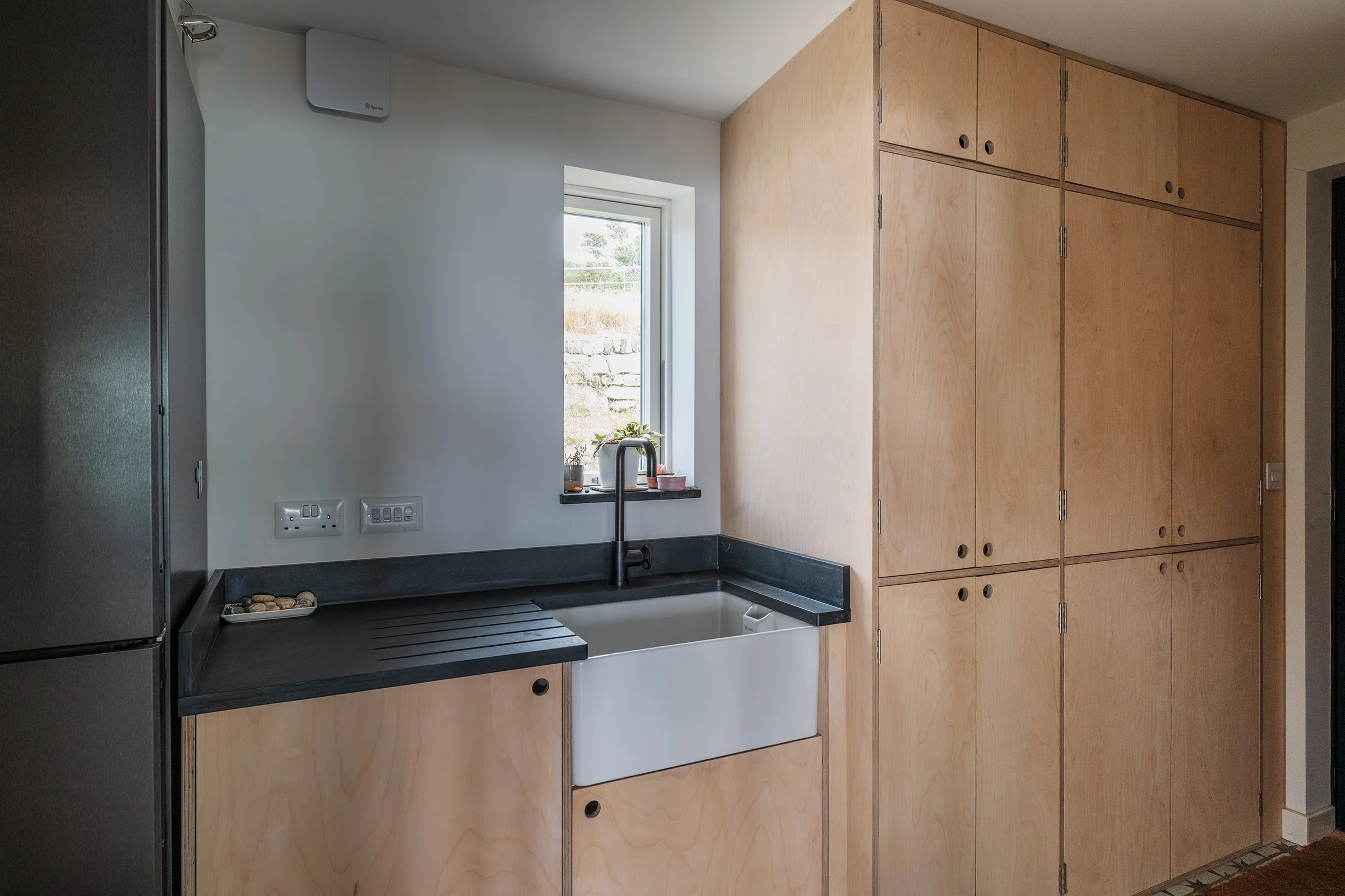Modern Utility Room with black countertops, farmhouse sink, wooden cabinets, refrigerator, and window with potted plants