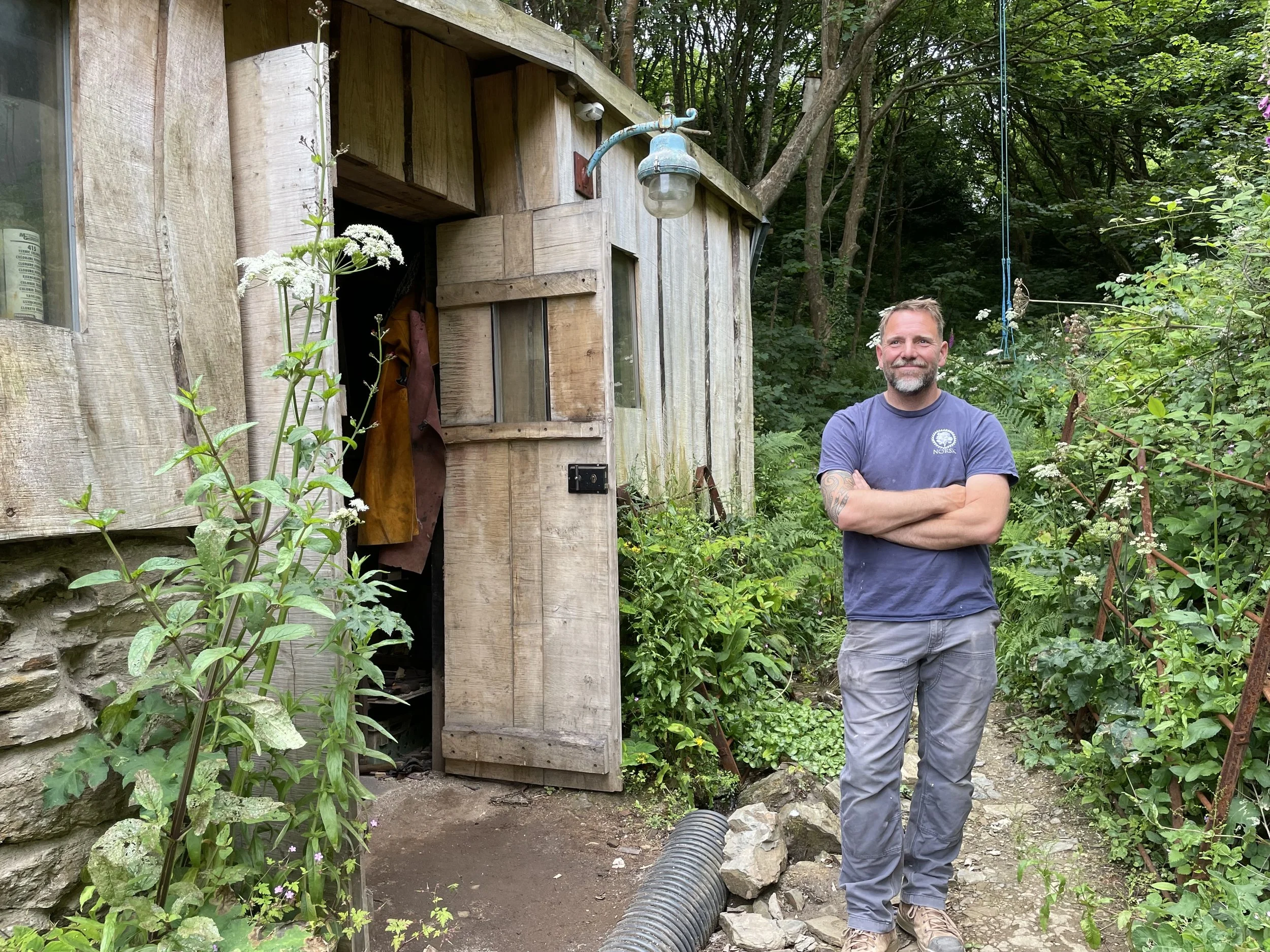 Dan Pye Carpenter and Norsk Founder with folded arms in front of a small wooden shed surrounded by greenery and flowers.