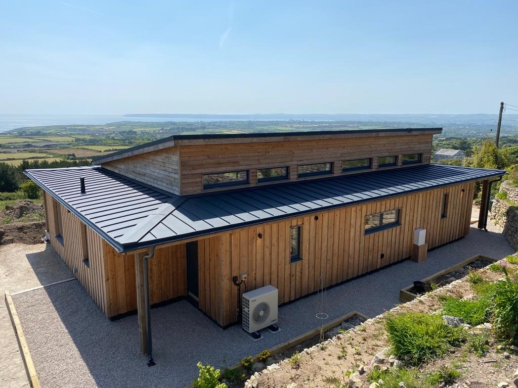 A modern wooden house on a hill with a metal roof, multiple windows, and a scenic view of the landscape and water in the background.