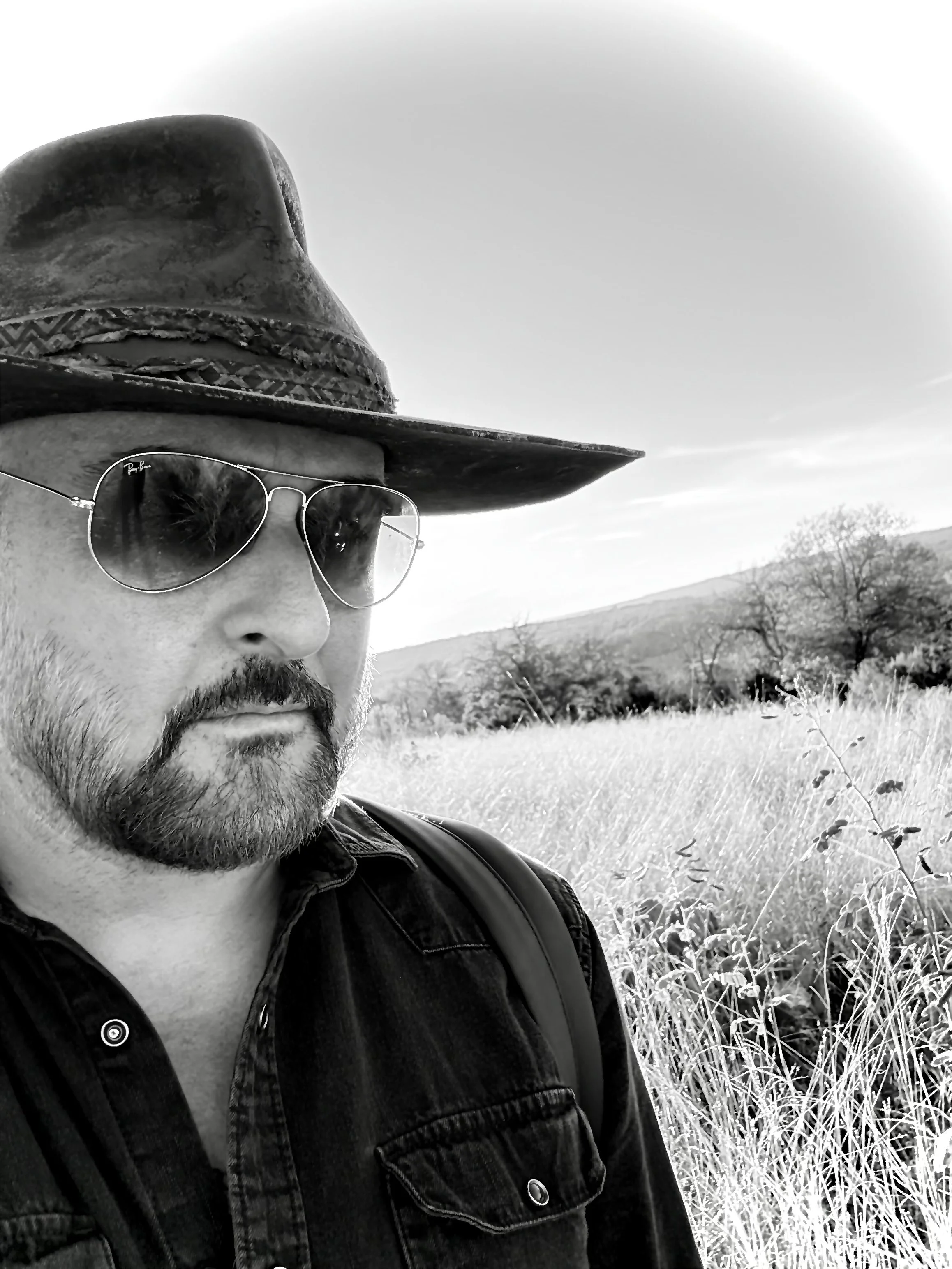 Close-up of a man wearing sunglasses and a wide-brimmed hat, outdoors in a grassy field with trees and hills in the background.