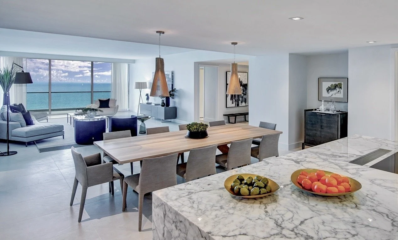 Open-concept living and dining area with large window view of the ocean, modern furniture, marble kitchen island with bowls of fruit