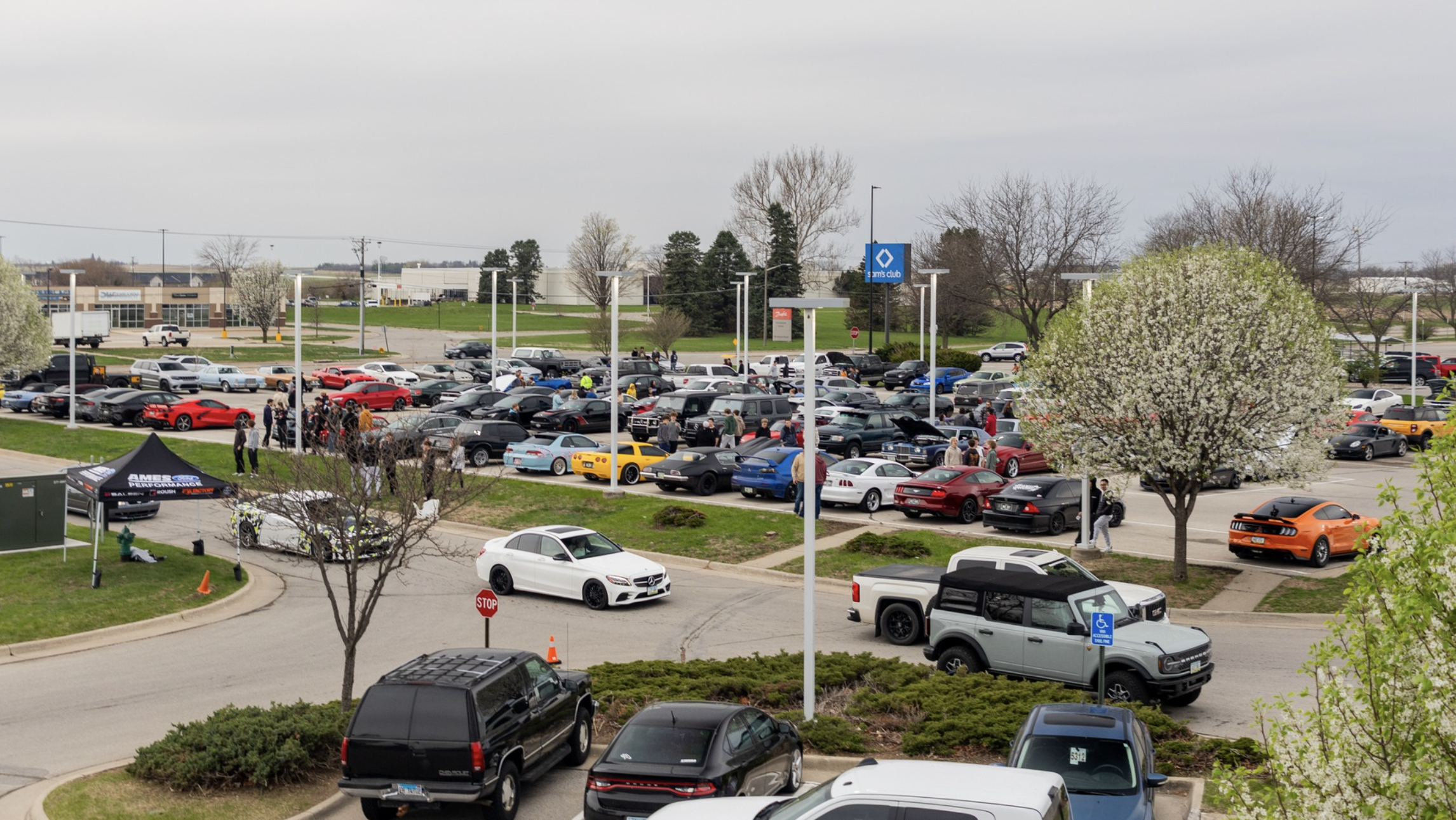 A parking lot filled with numerous cars, and people walking around, possibly at a car event or gathering.