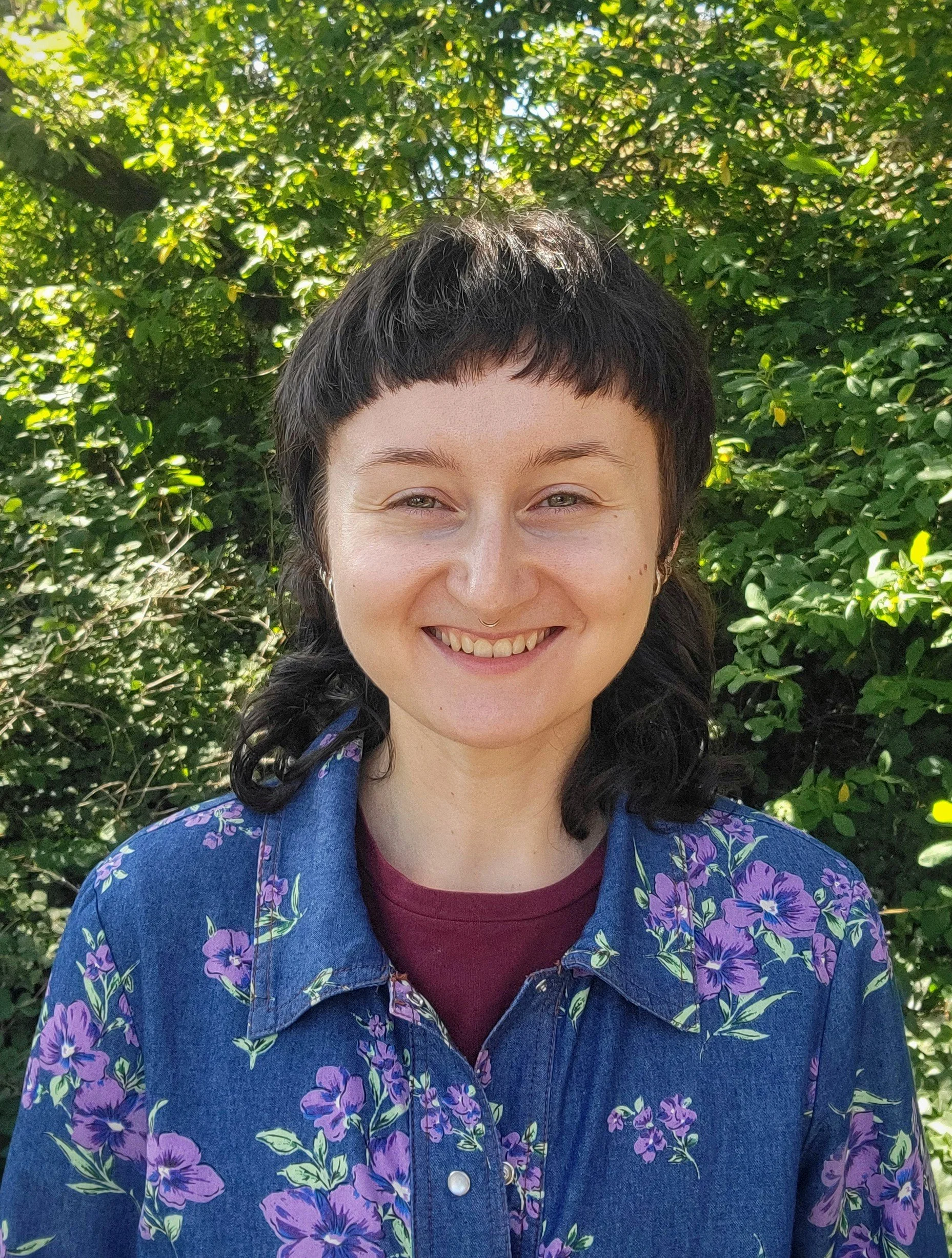 A woman with short dark hair, wearing a floral denim jacket and a maroon shirt, smiling outdoors in front of green leafy bushes.
