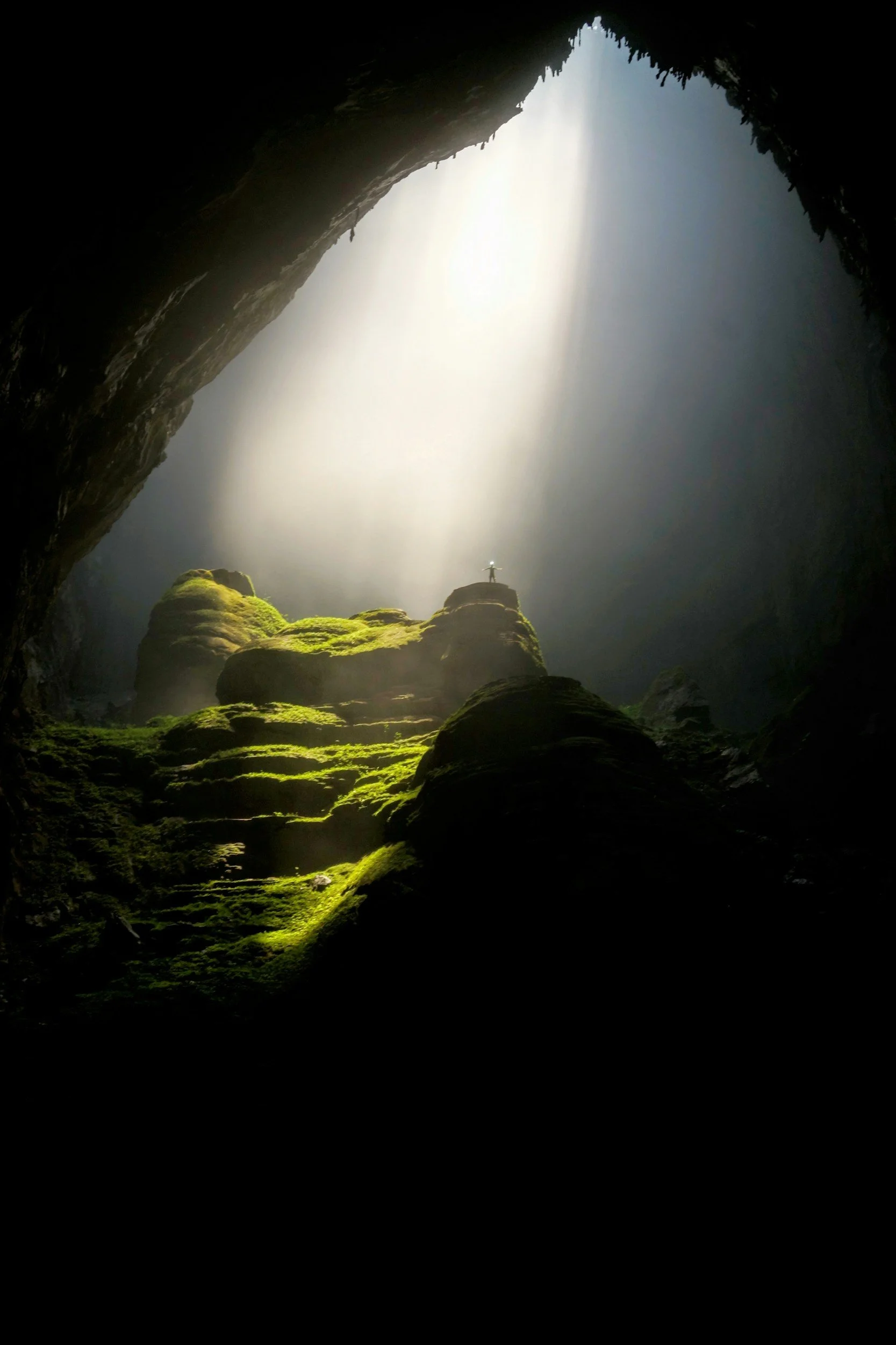 A person standing on moss-covered rocks inside a large cave with sunlight streaming down from an opening above.