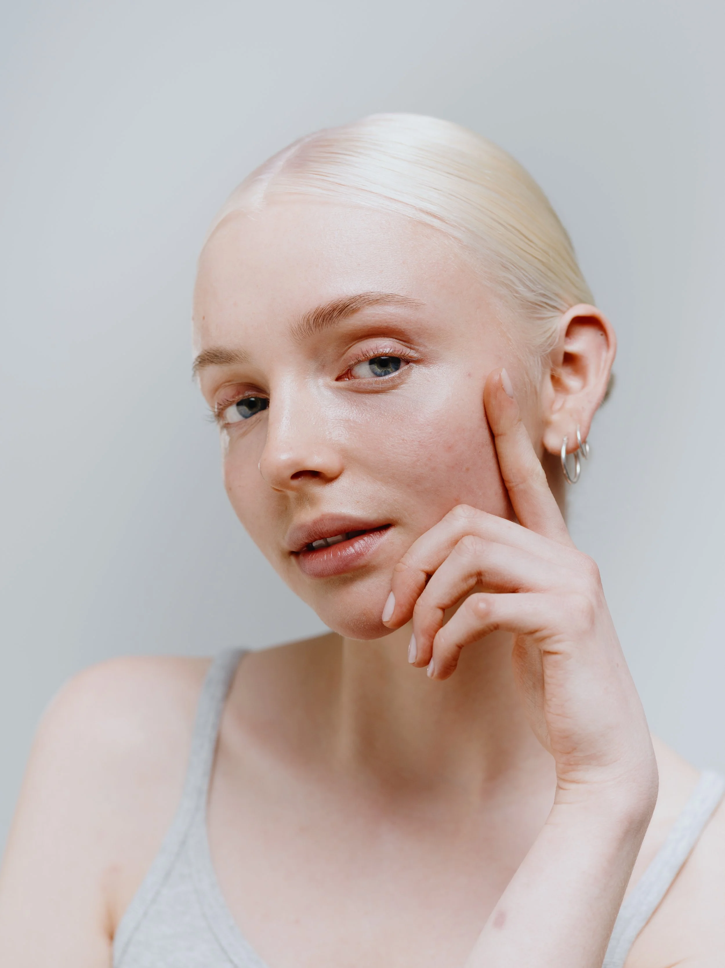 Nahaufnahme eines jungen Mädchens mit blauer Augenfarbe, blonder Haut und einer grauen Trägerleibchen, das mit der Hand an ihrem Gesicht berührt, vor einem hellen Hintergrund.