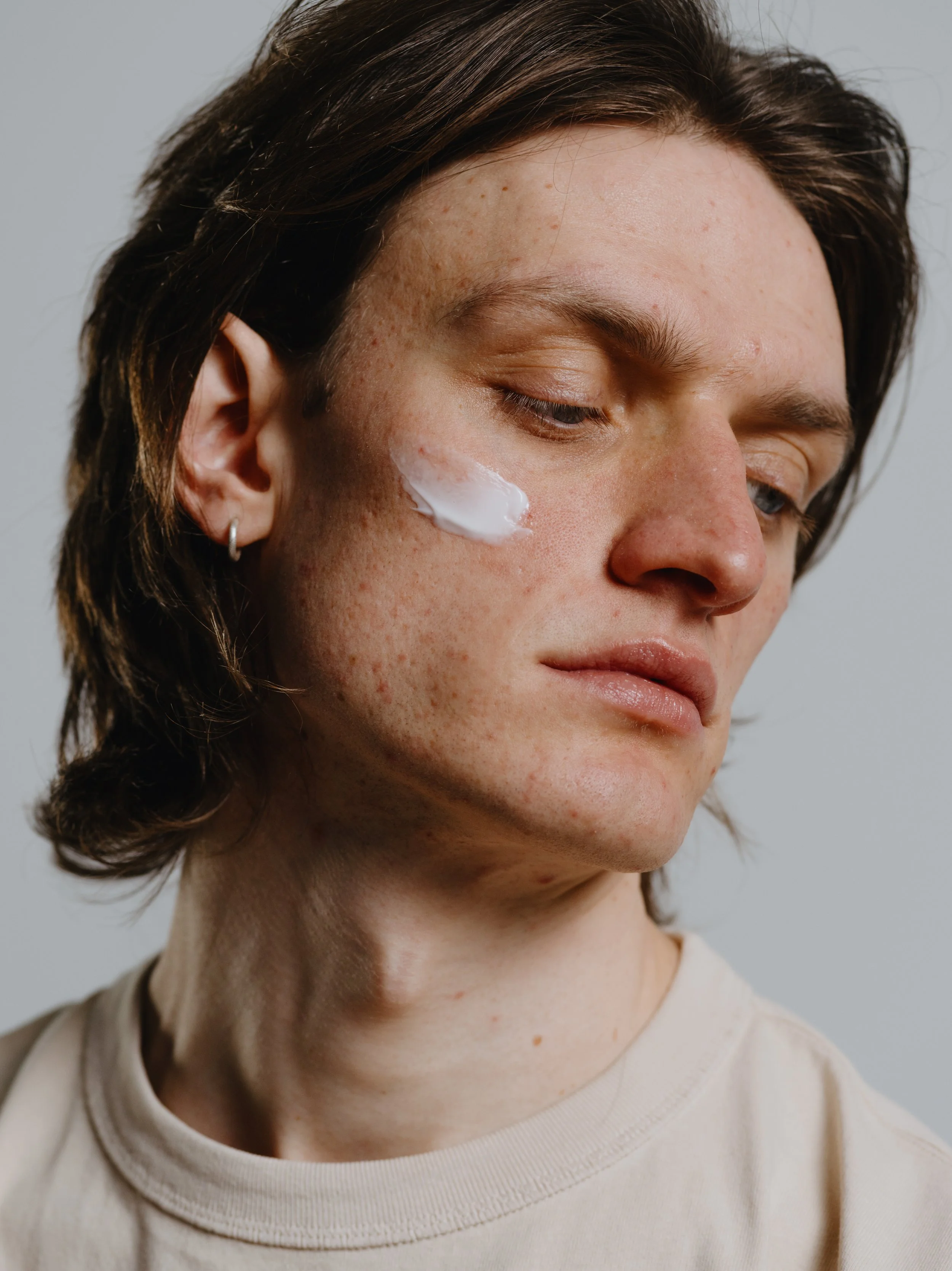 Nahaufnahme eines jungen Mannes mit braunen Haaren, der die Augen geschlossen hat. Auf der Wange trägt er eine weiße Creme. Er trägt ein weißes T-Shirt.