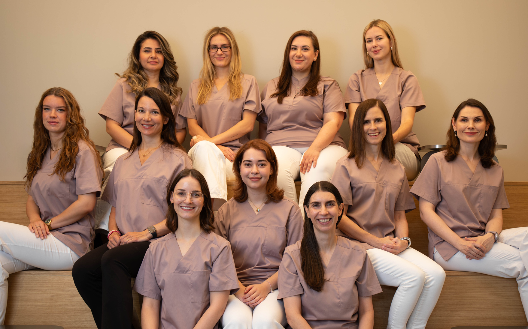 Eine Gruppe von Frauen in braunen medizinischen Uniformen sitzt und steht auf einer Treppe in einem Raum mit neutraler Wandfarbe, alle lächeln.