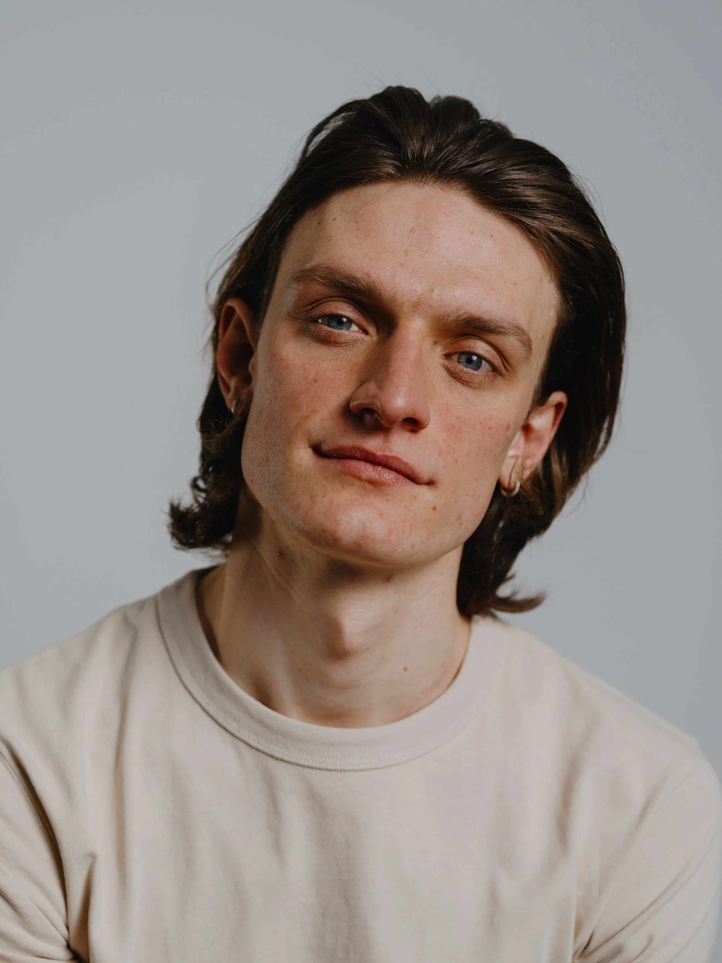 Nahaufnahme eines jungen Mannes mit mittellangen dunklen Haaren, blauen Augen und heller Haut, trägt ein cremefarbenes T-Shirt, vor hellem grauem Hintergrund.