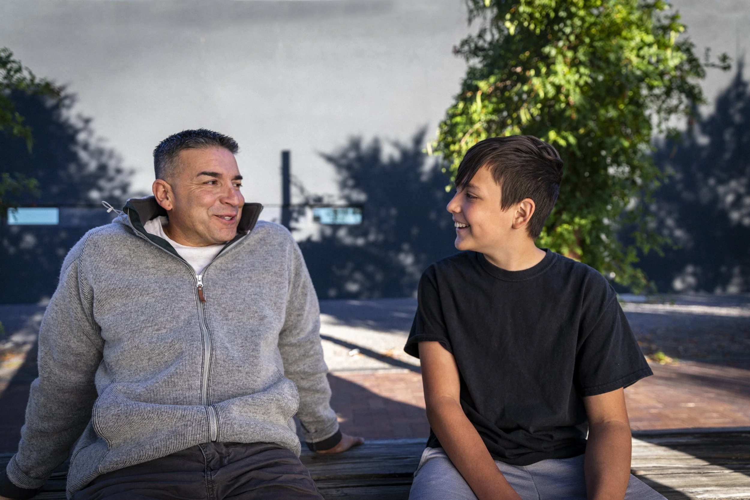 A man and a boy sitting on a park bench, smiling and looking at each other, with trees in the background.