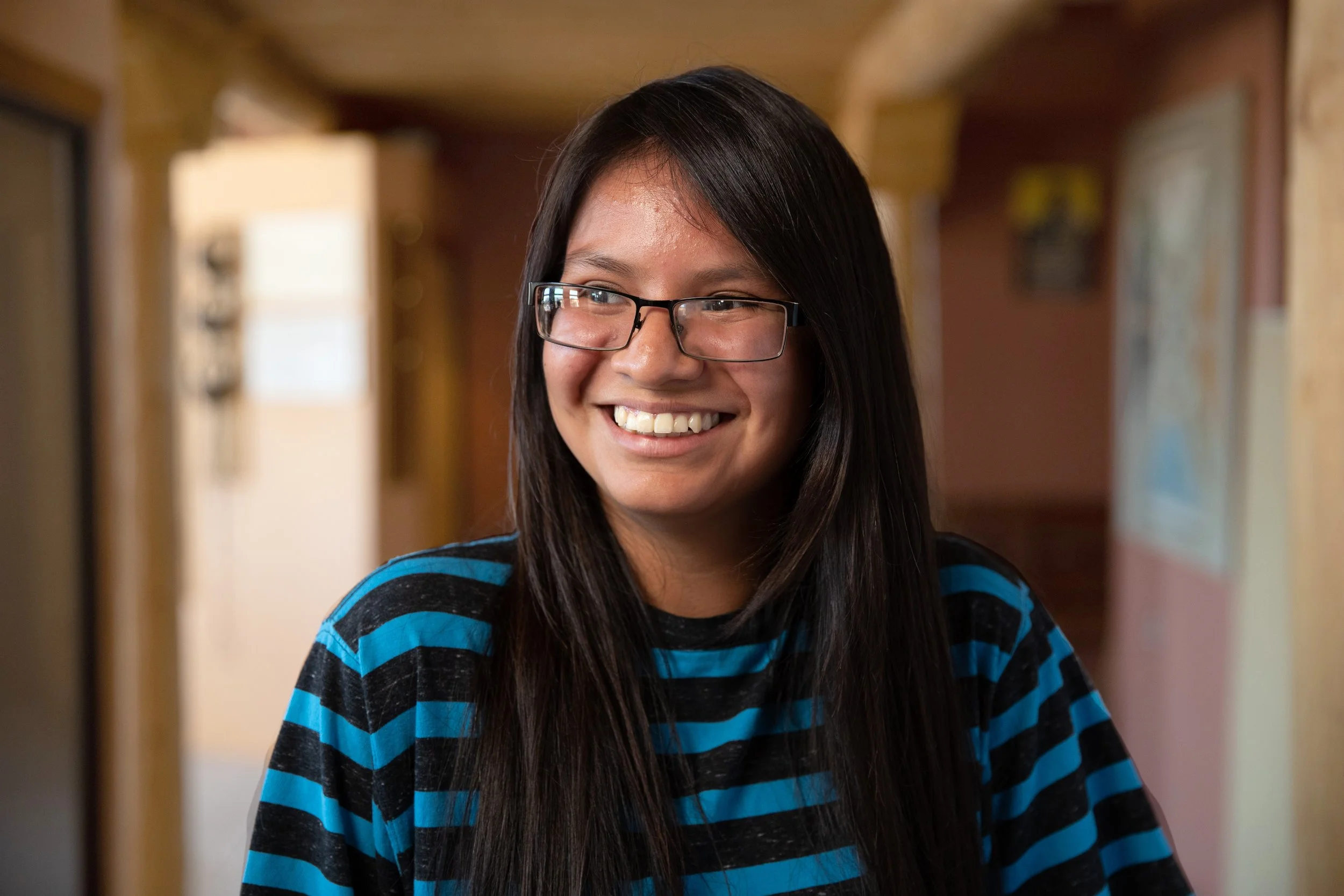 A young woman with long dark hair and glasses smiling in a hallway, wearing a black and blue striped shirt.