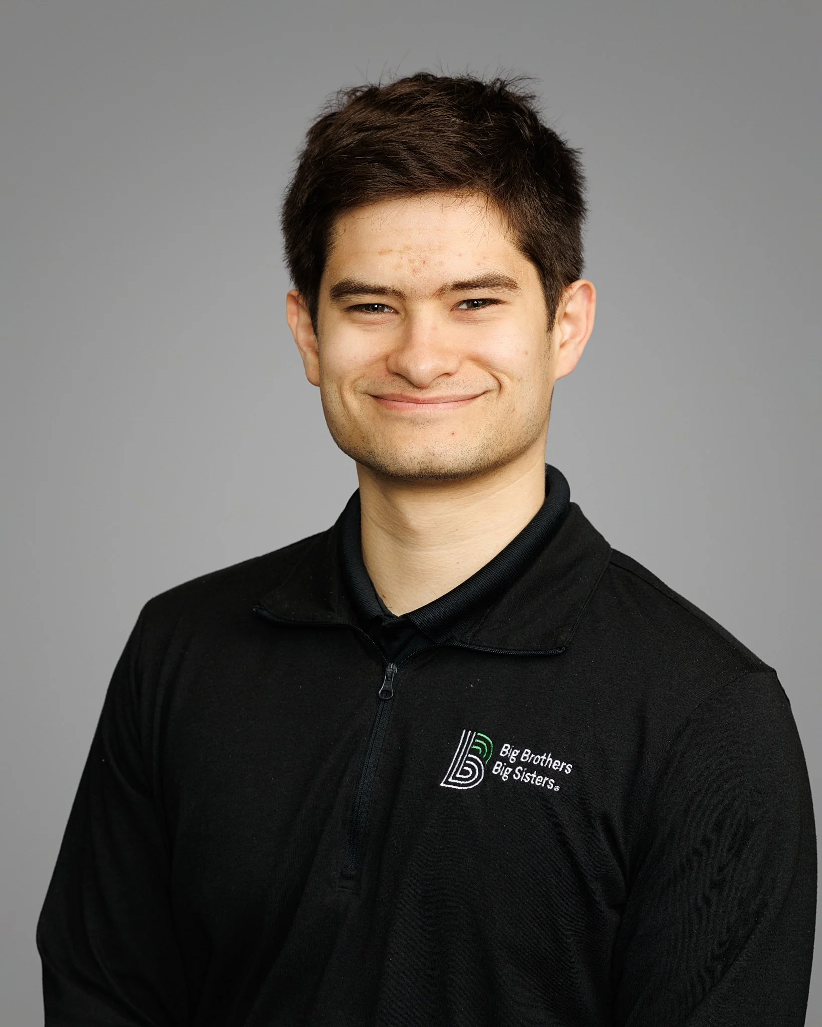 Young man smiling in front of a "Big Brothers Big Sisters of Washington" logo on a dark wall. The atmosphere is positive and welcoming.