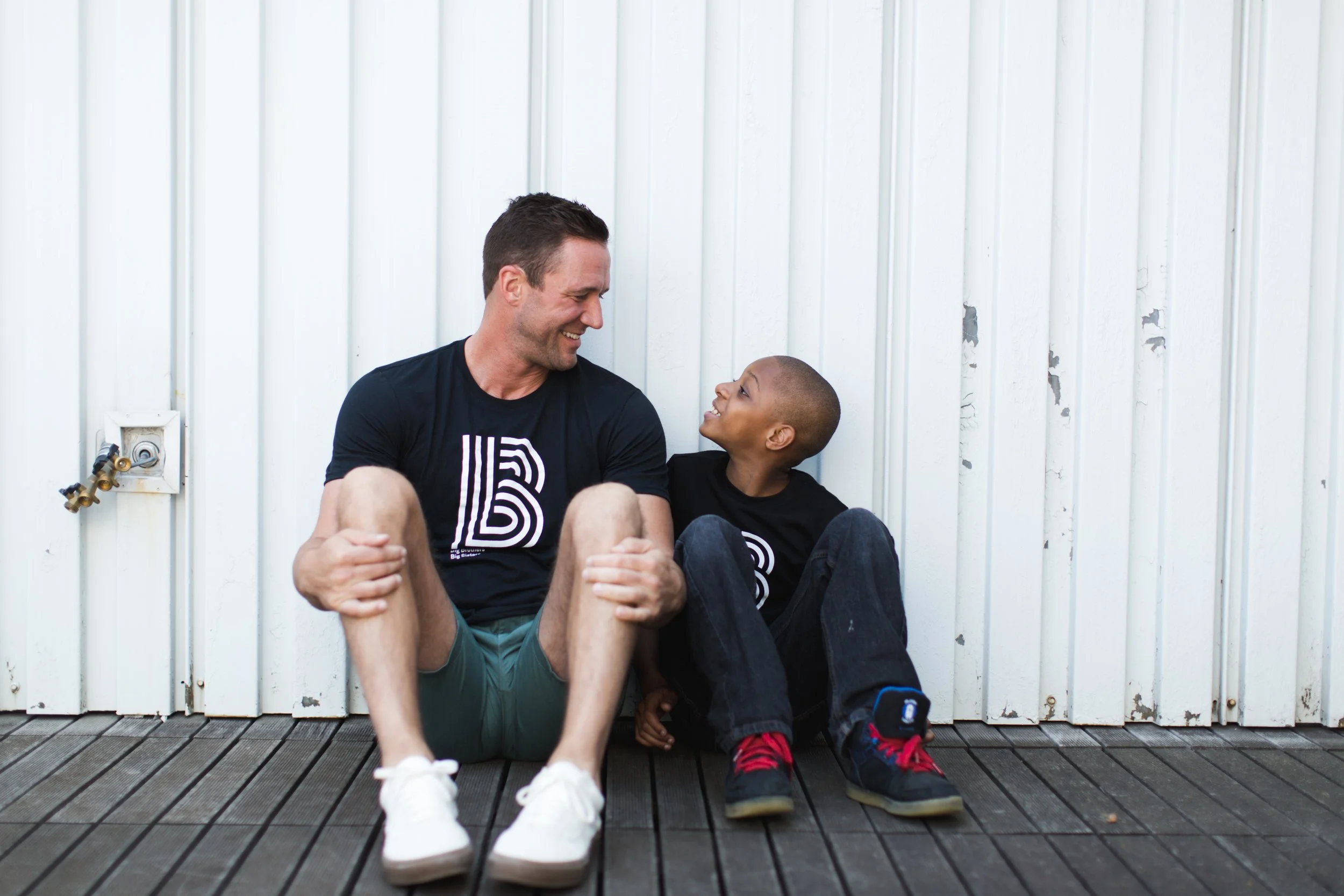 A man and a boy sitting on a wooden deck against a white wall, smiling and looking at each other.