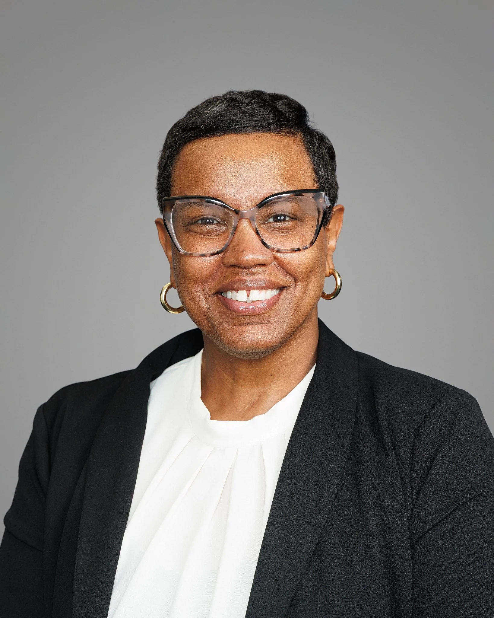 A woman with braided hair and glasses smiles warmly. She wears a navy blue zip-up jacket. The background features a light patterned wall, conveying a friendly and relaxed atmosphere.