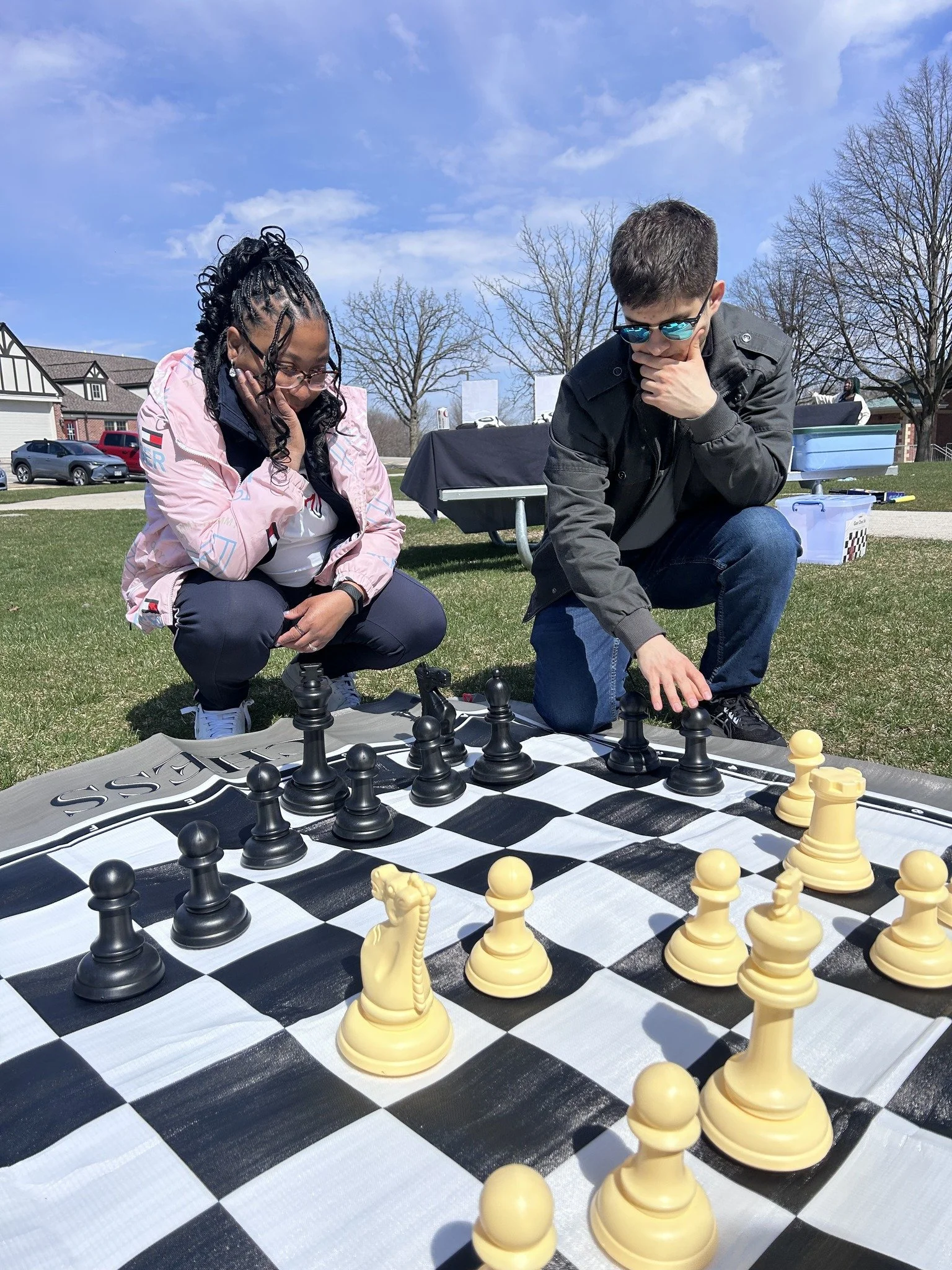 Two people are playing chess outdoors on a large black-and-white checkered chessboard on the grass, with a picnic table and trees in the background, under a blue sky with some clouds.