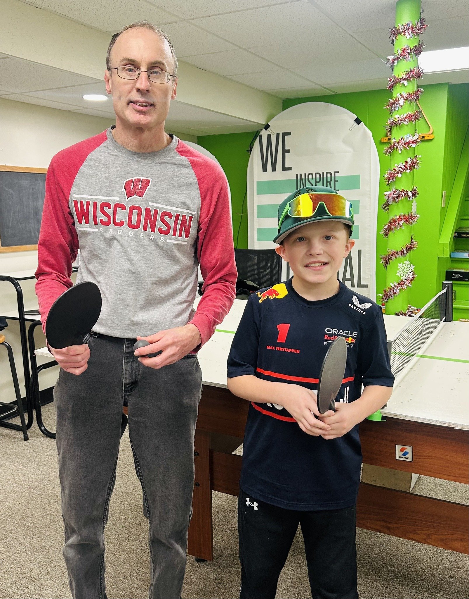 A man and a young boy smile while holding ping pong paddles in a recreation room. The background features a motivational banner and green walls.