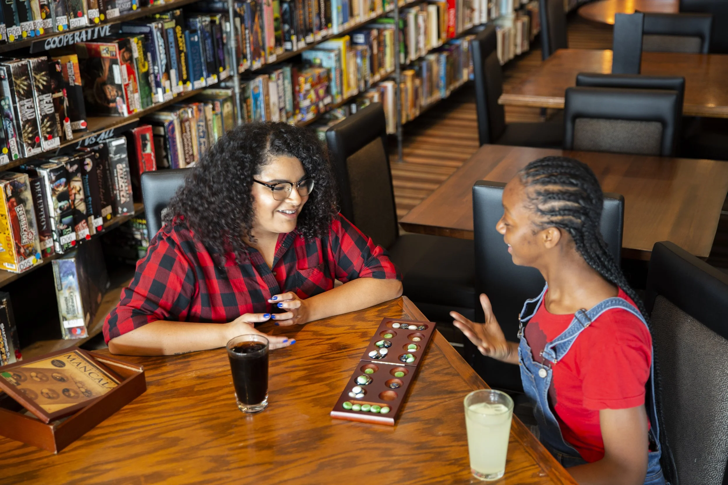Two women sitting at a wooden table in a library, playing a board game with colorful marbles, surrounded by books on shelves.