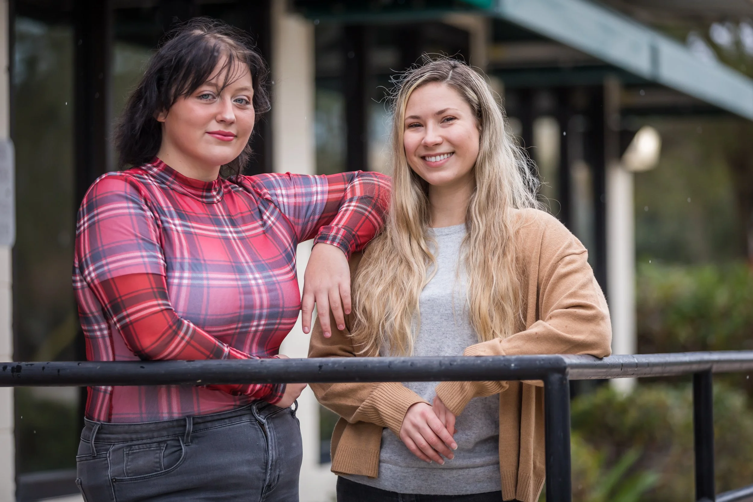 Two women standing outdoors near a railing, with one woman in a red plaid shirt and the other in a tan jacket, smiling at the camera.