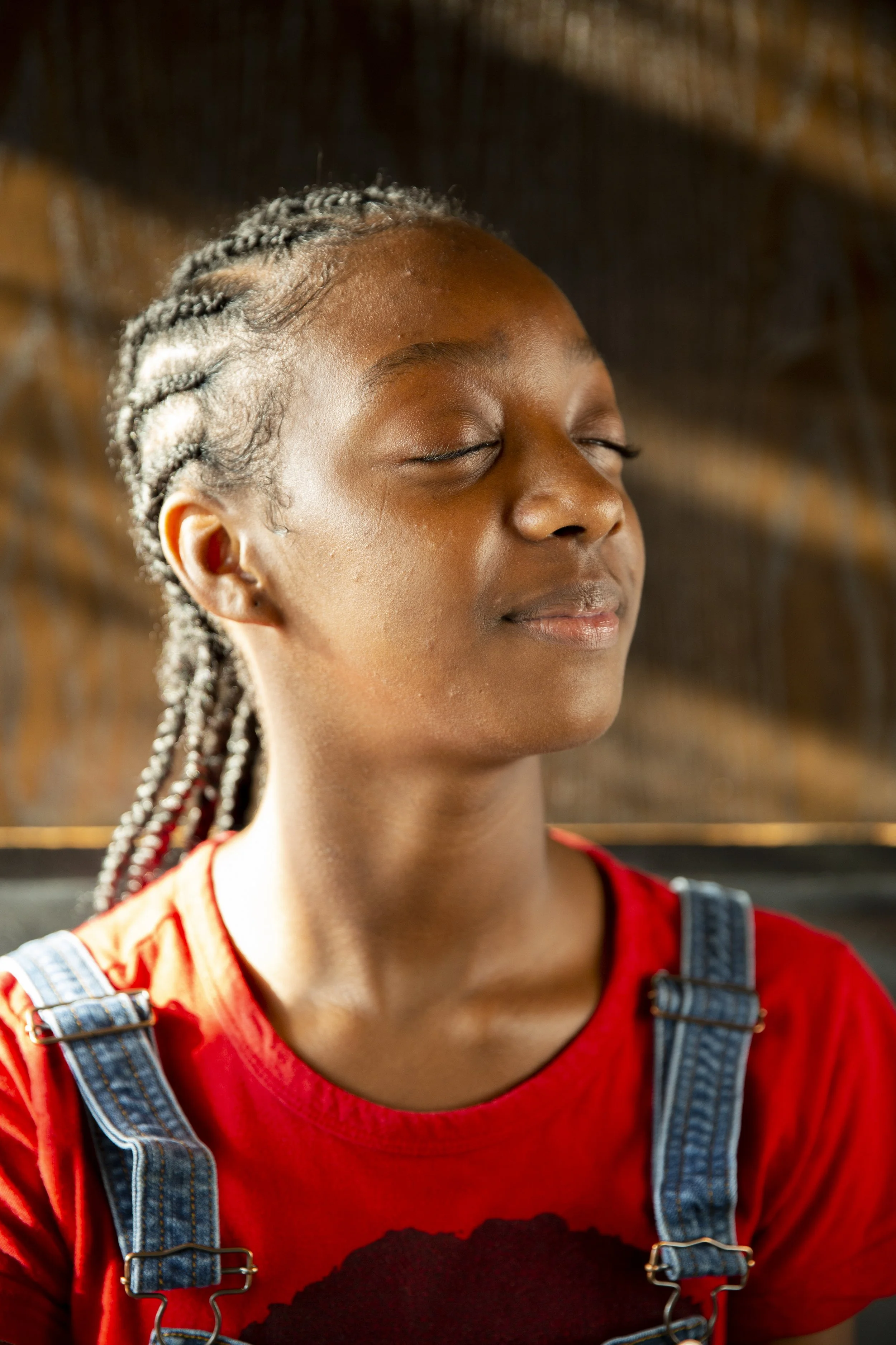 A young woman with braided hair and closed eyes stands outside. She wears a red shirt with a black design and denim overalls. The background appears to be natural with a blurred brownish setting.