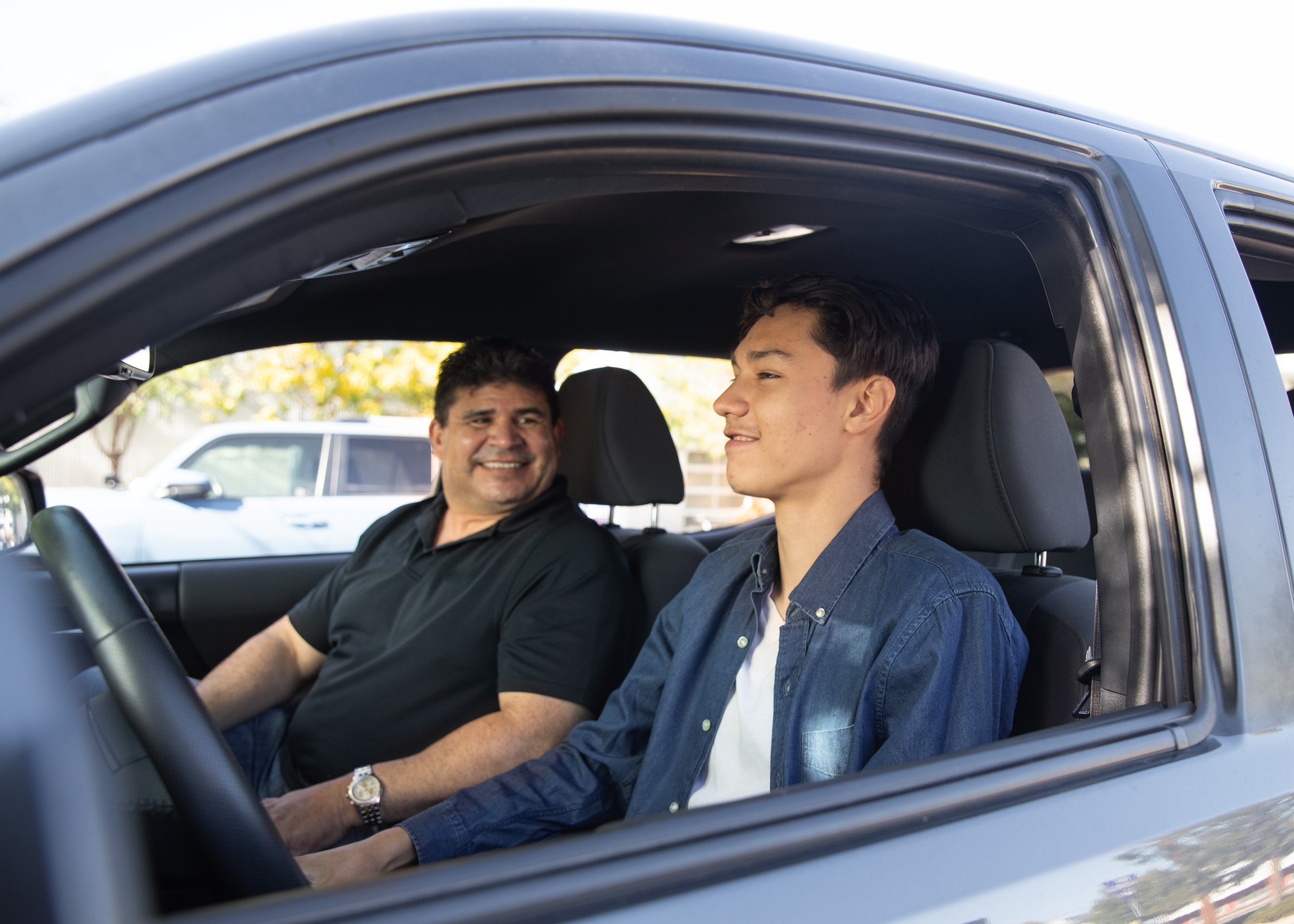 A man and a teenage boy sitting inside a car, smiling at each other, with trees and parked cars visible outside through the open window.
