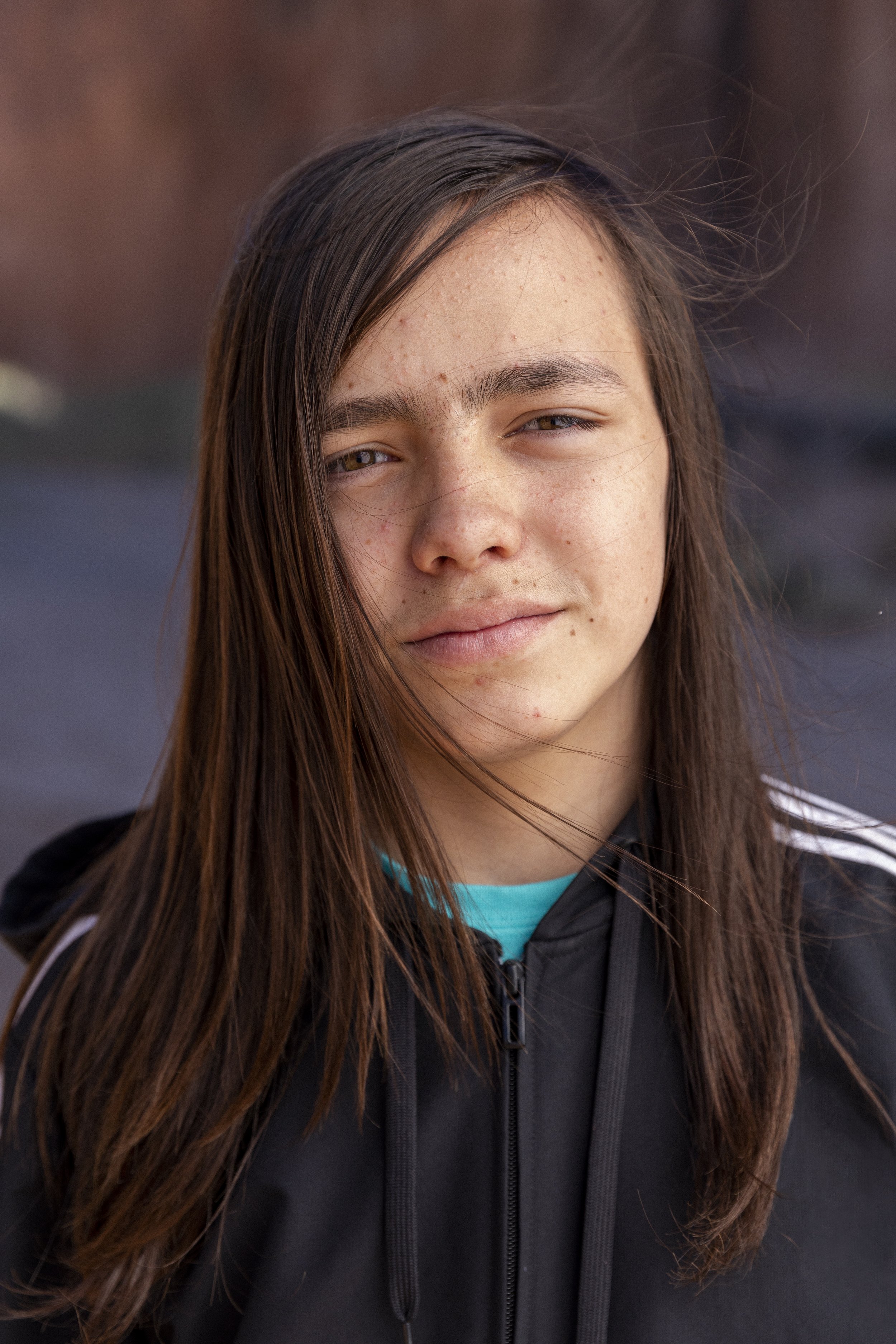 A young person with long brown hair, slightly smiling, wearing a black hoodie with a turquoise shirt underneath, outdoors on a cloudy day.