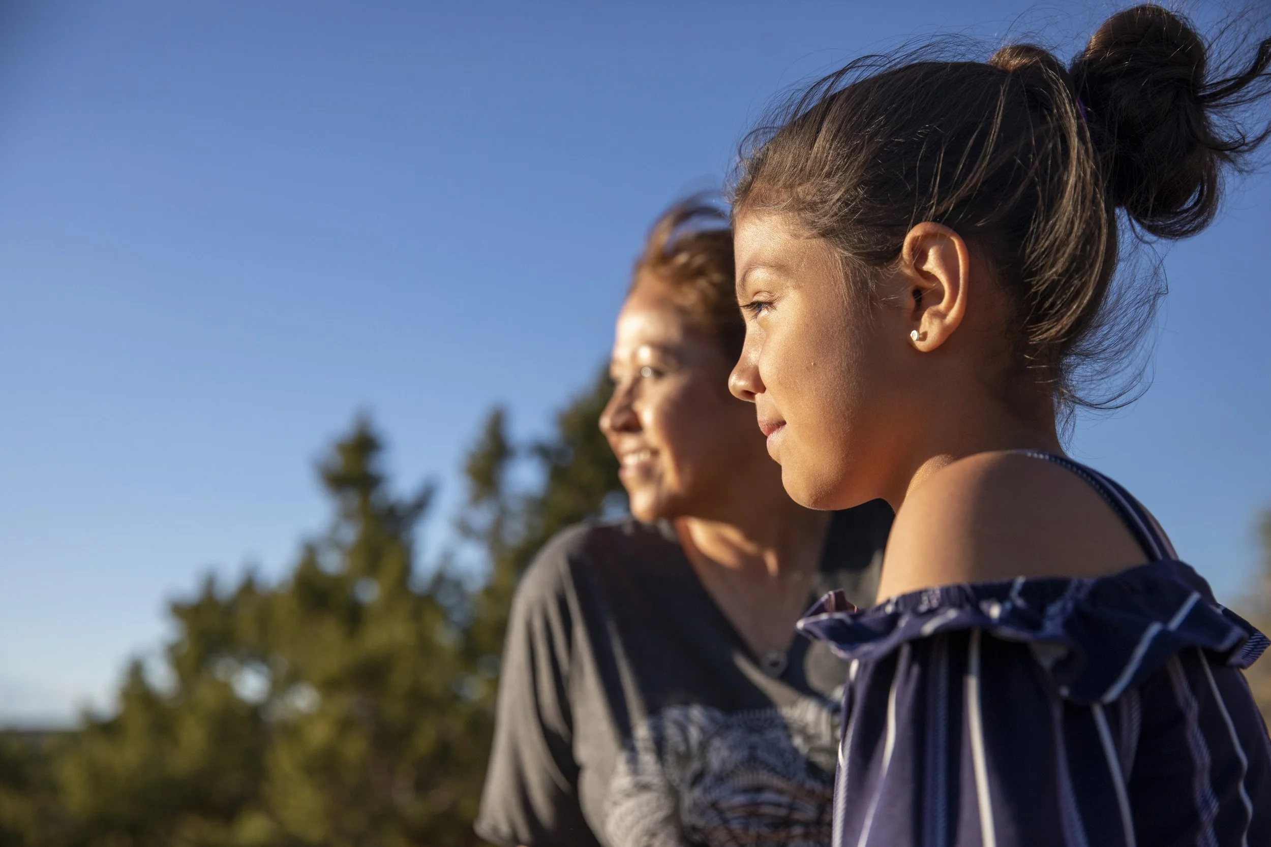 A young girl and an older woman gaze thoughtfully into the distance against a clear blue sky, surrounded by blurred green trees, conveying serenity.
