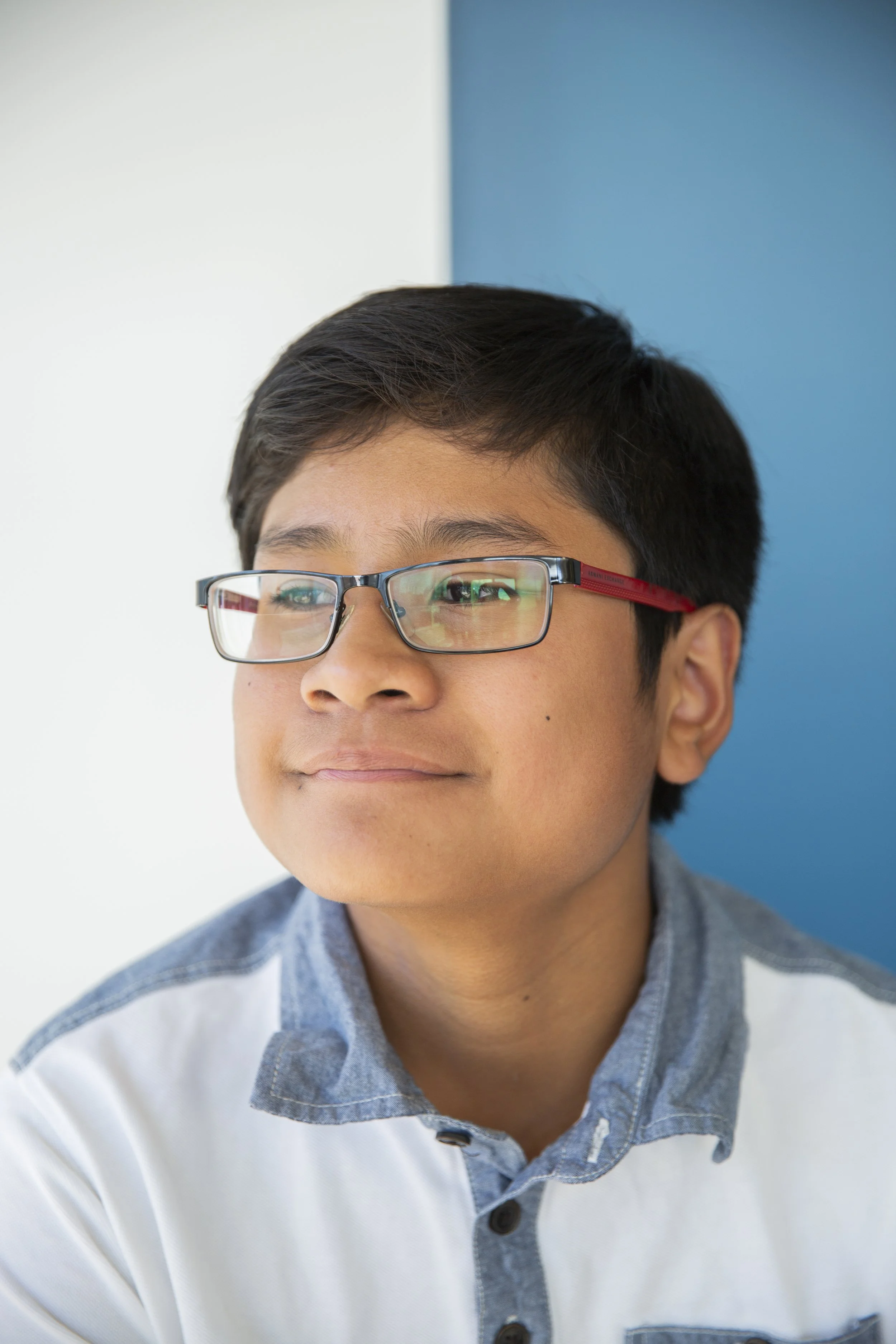 Close-up of a young boy with glasses, wearing a collared shirt and a sweater, against a background divided in white and blue color.