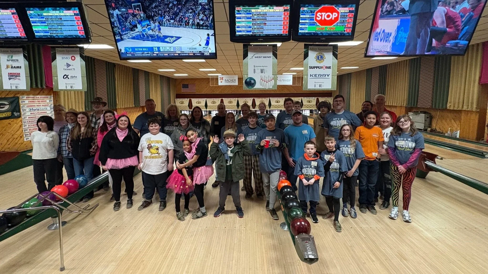 A group of people, including children and adults, gather happily in a bowling alley. Brightly colored balls are visible, and TV screens show sports above.