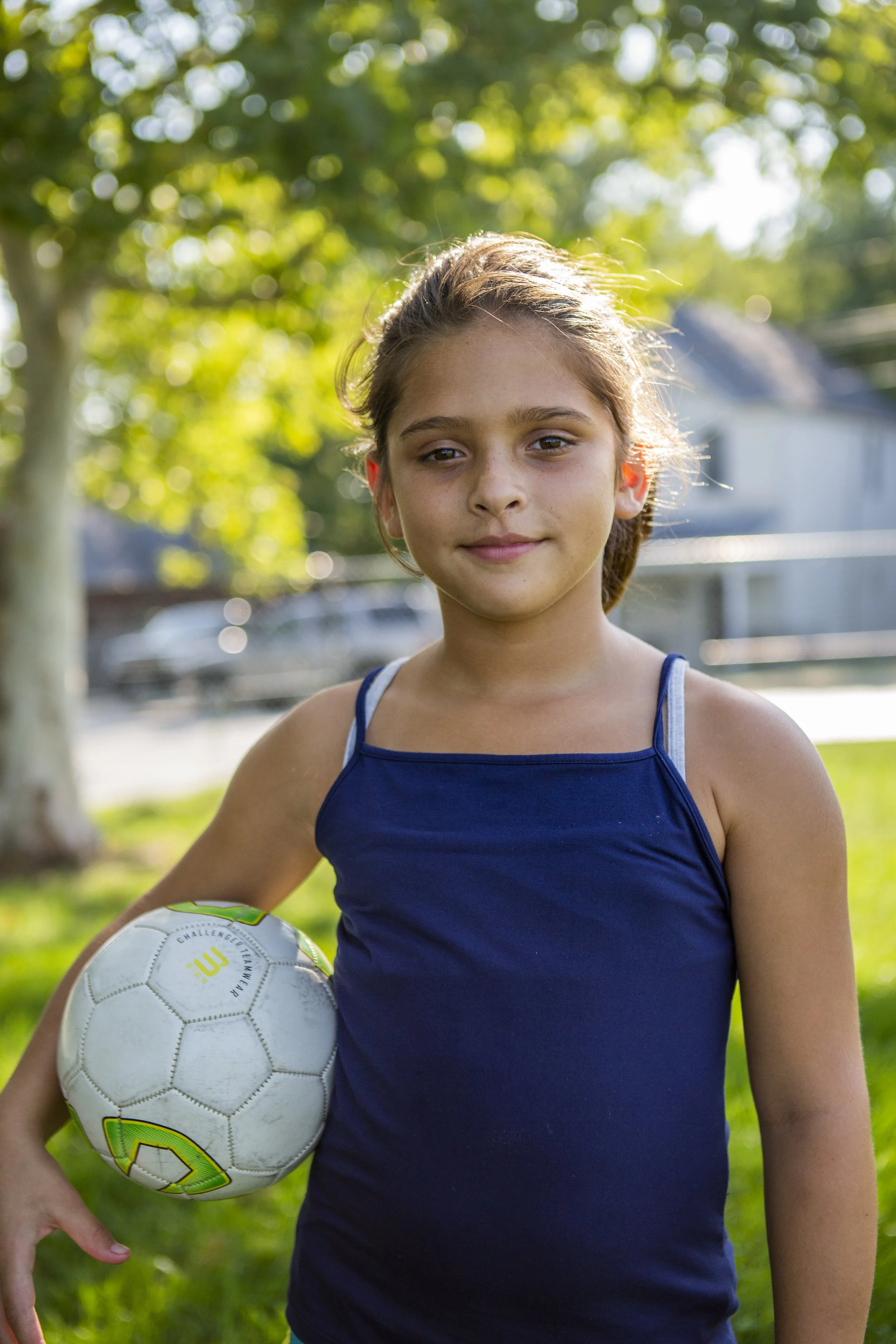 A young girl stands on grass, holding a soccer ball under her arm. She's wearing a navy tank top, with sunlight filtering through trees in the background.